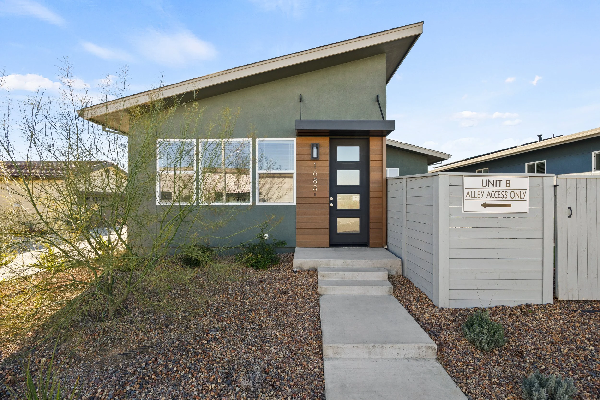 Front view of a modern green house with a sloped roof, black front door with four glass panels, and a small concrete staircase leading to the entrance. There is a white fence on the right with a sign indicating "Unit B Alley Access Only." Desert land