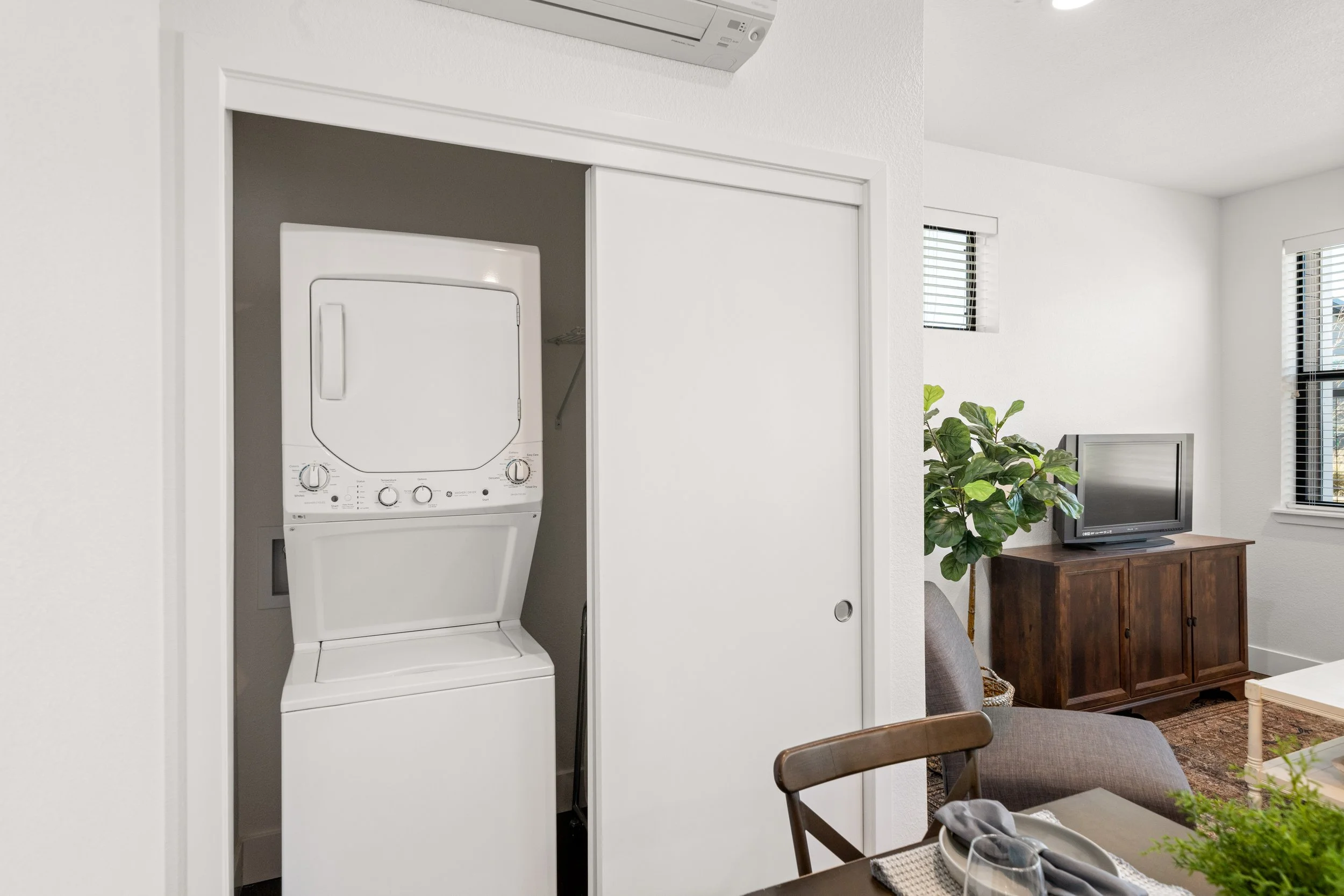 Small laundry area with a stacked white washer and dryer inside a closet with a sliding door, adjacent to a living room with a television on a wooden stand, a large plant, and windows with blinds.