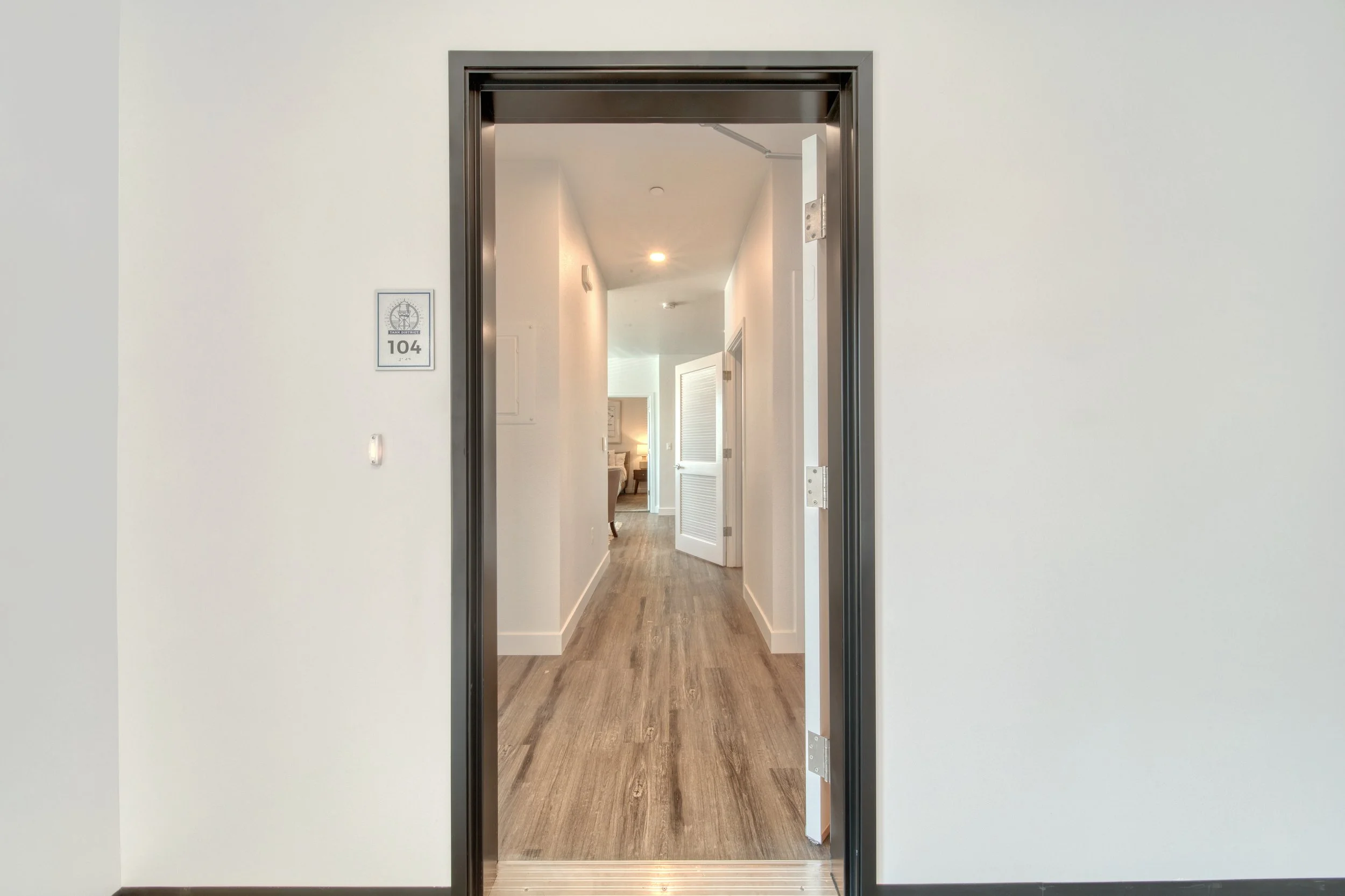 View through an elevator door into a hall with wooden flooring and white walls, with some furniture visible at the end.