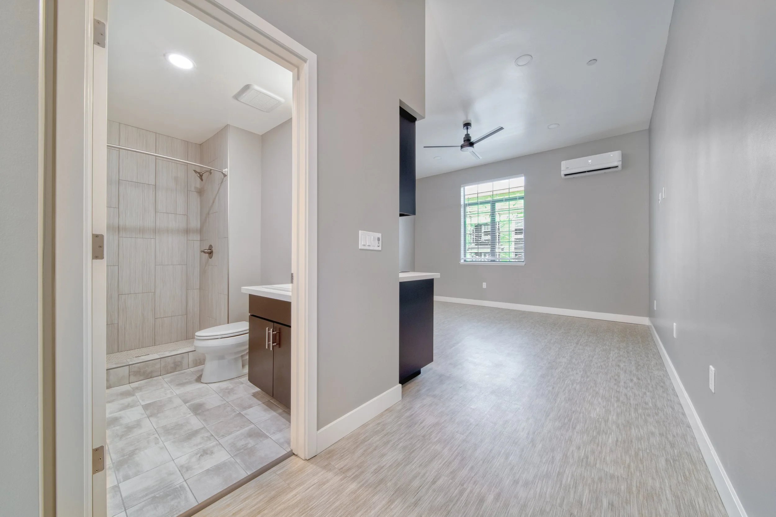 Empty room with gray walls, a window, ceiling fan, and wall-mounted air conditioner. An adjacent bathroom with a shower, toilet, and vanity is visible.