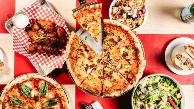 Pizza, fried chicken wings with dipping sauce, pasta salad, caesar salad, and breadsticks on a table.
