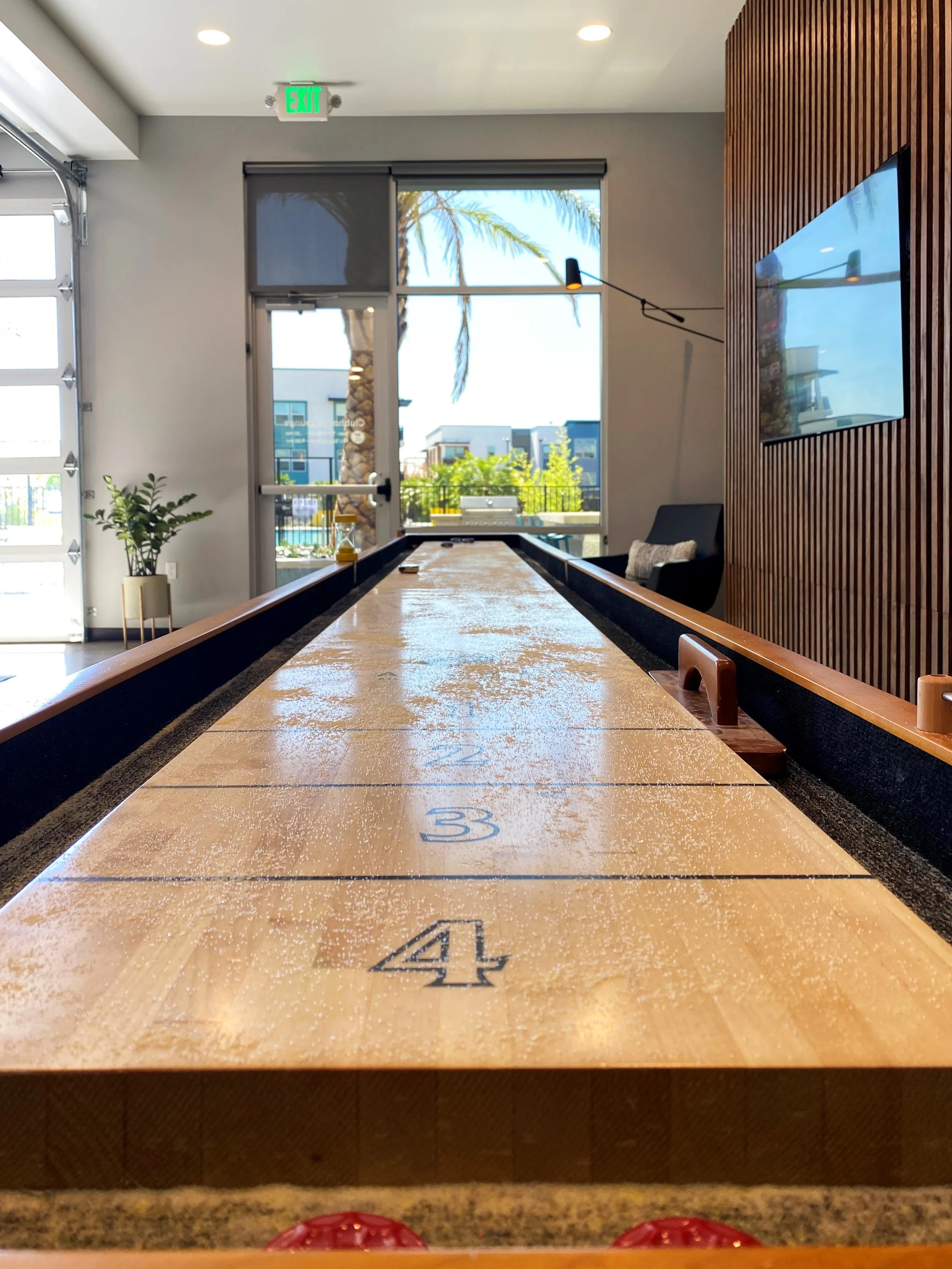 Indoor shuffleboard table with numbered scoring zones, surrounded by a modern lounge area with a television on a wooden-paneled wall and a large glass door showing outdoor palm trees.