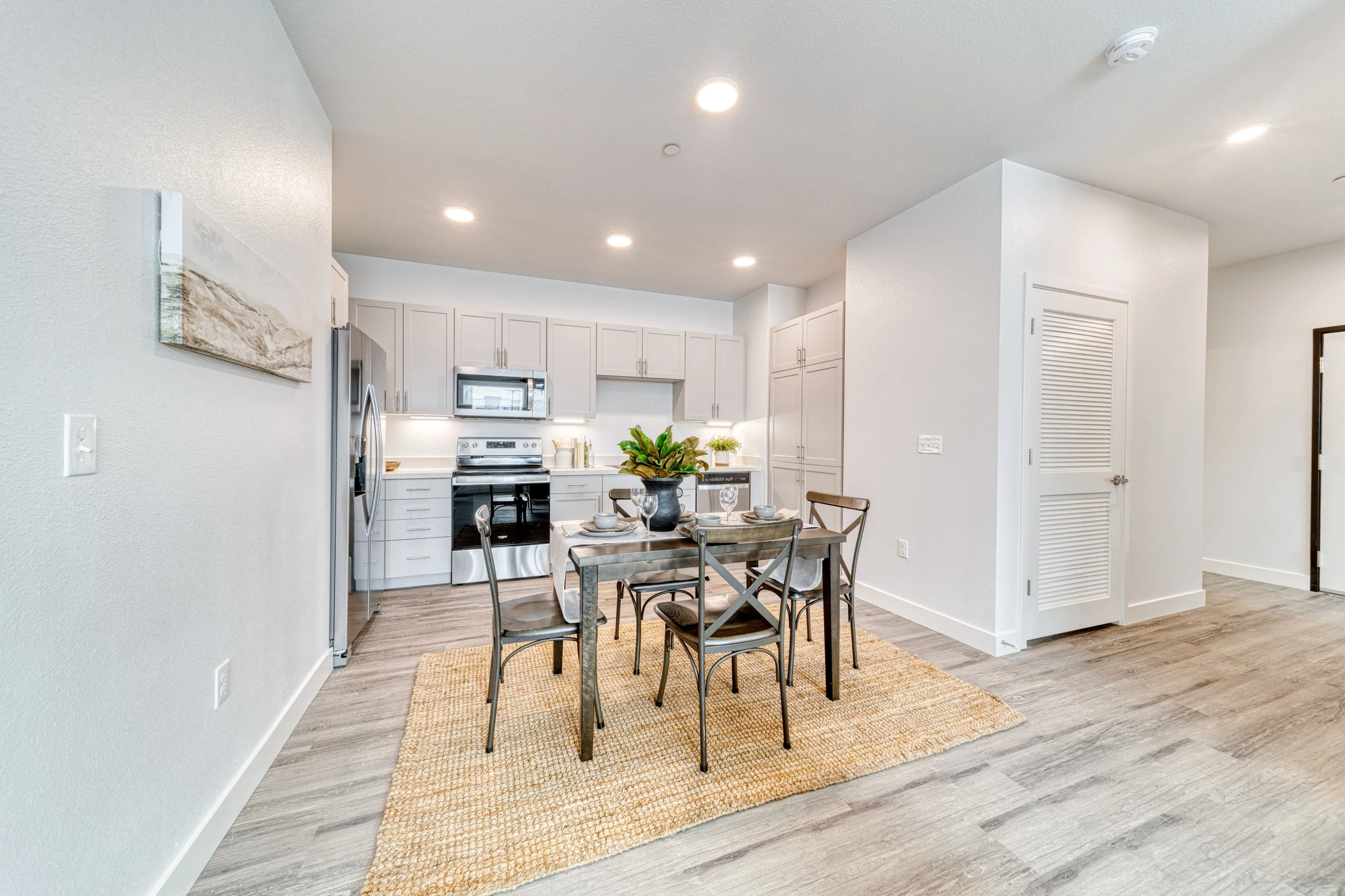 Dining area with a wooden table set for four, a potted plant centerpiece, on a woven rug, in front of a modern kitchen with white cabinets and stainless steel appliances.