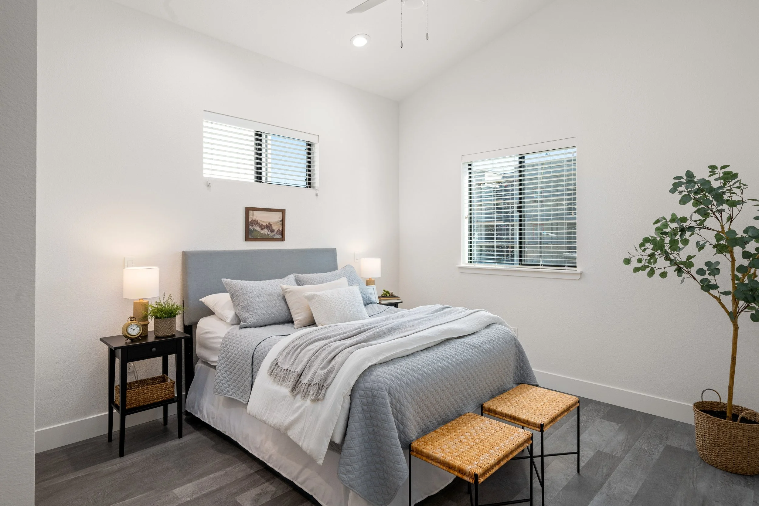 A minimalist bedroom featuring a bed with gray quilted bedding and multiple pillows, two bedside tables with lamps, a small framed artwork above the bed, a potted plant in a woven basket, and two woven stools at the foot of the bed, with three window