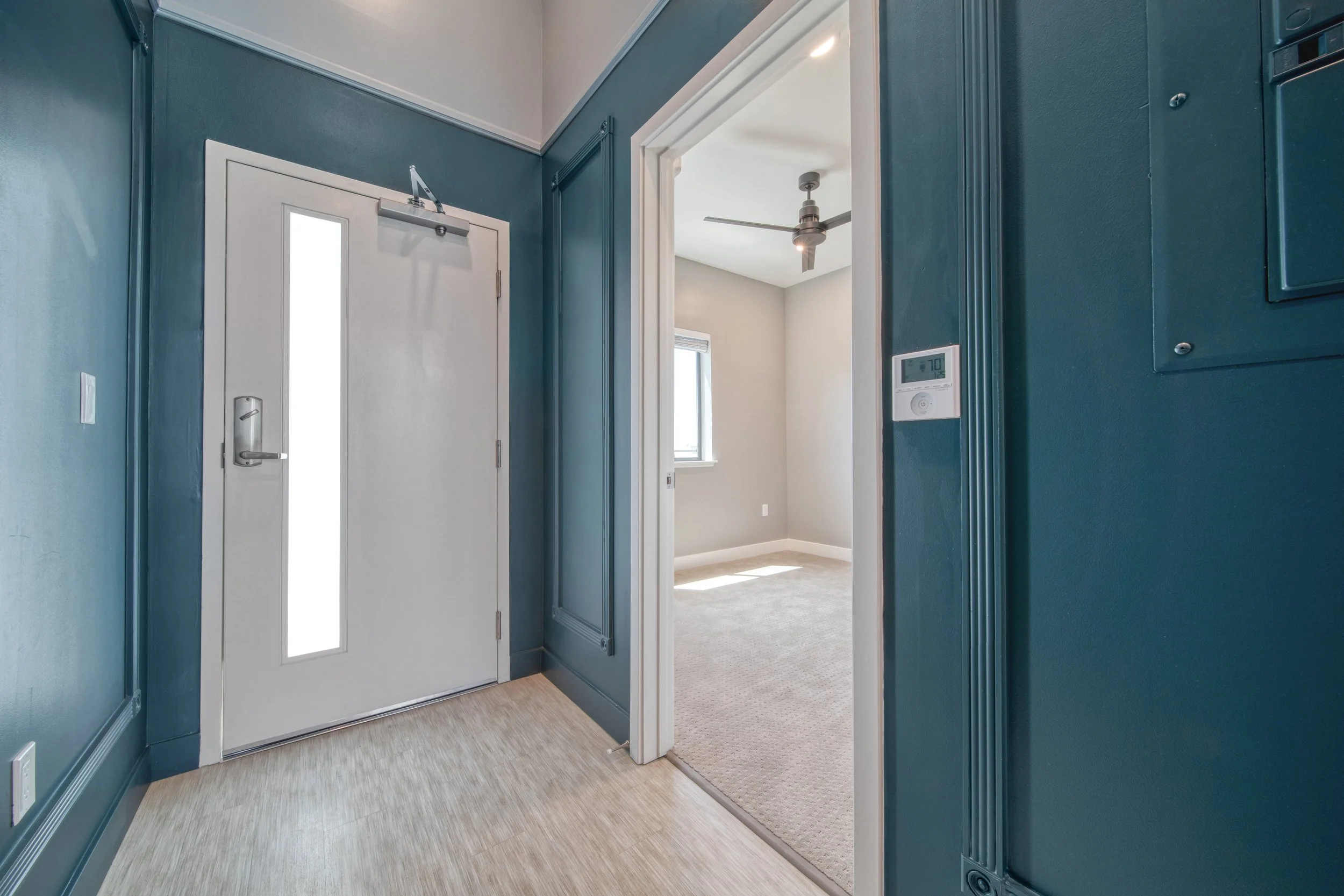 Entryway with white front door and adjacent light gray carpeted room, blue walls, ceiling fan, and thermostat on wall.