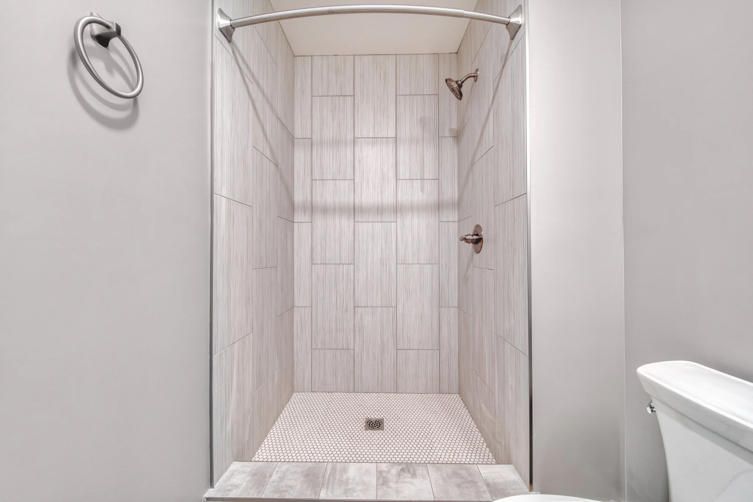 Empty walk-in shower with tiled walls, a showerhead, and a drain, adjacent to a toilet, with a simple gray wall and towel ring on the side.