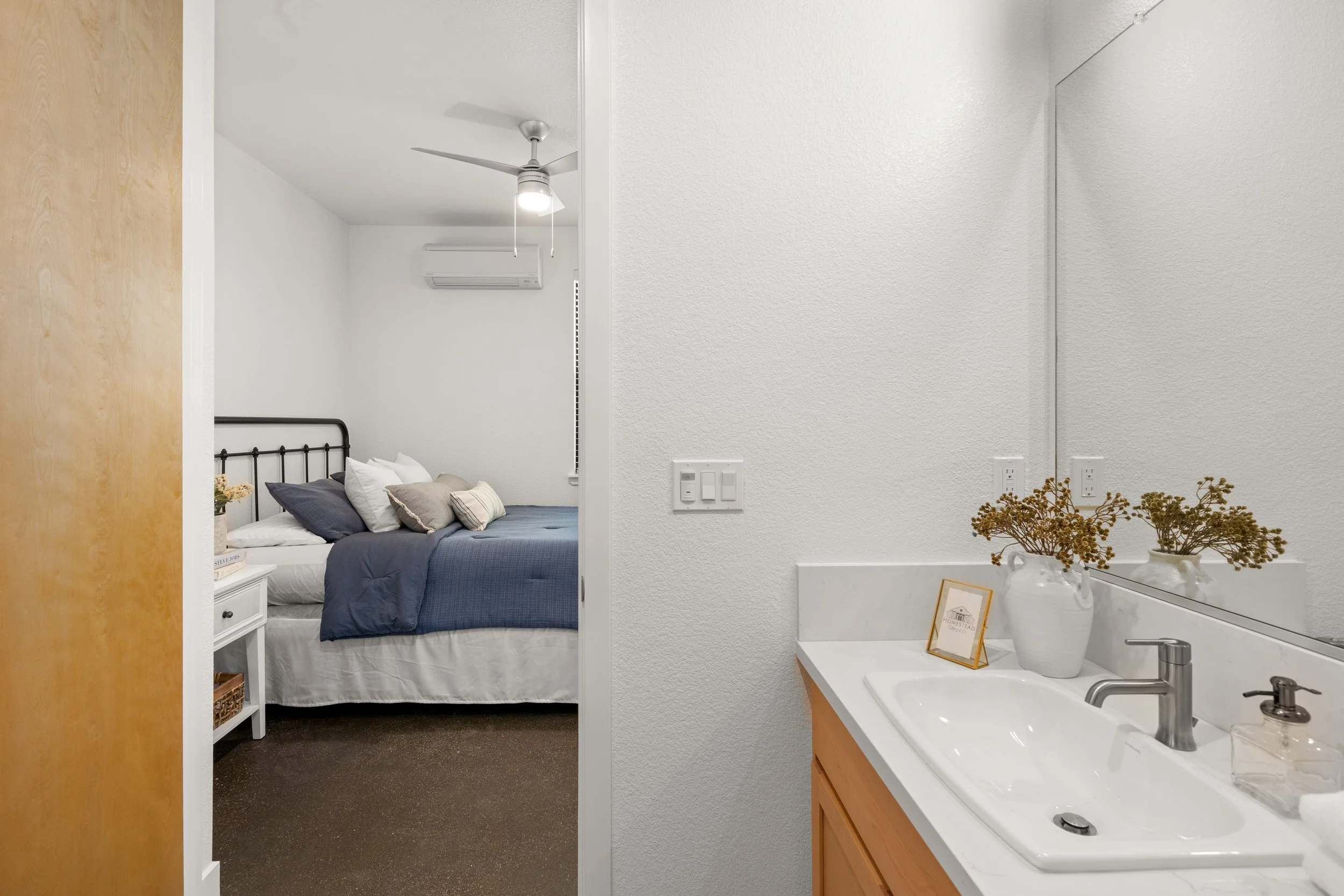 View of a bedroom with a white wall, blue bedding, and throw pillows, seen from a bathroom with a white sink and mirror, a vase of dried flowers, and a small framed picture.