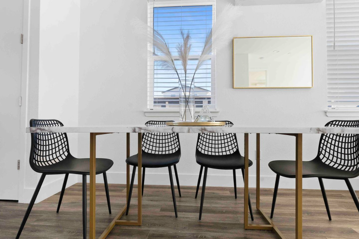 Modern dining room with a marble-top table, five black grid-back chairs, a vase with pampas grass, and a rectangular mirror on the white wall. There is a window with blinds showing a blue sky outside.