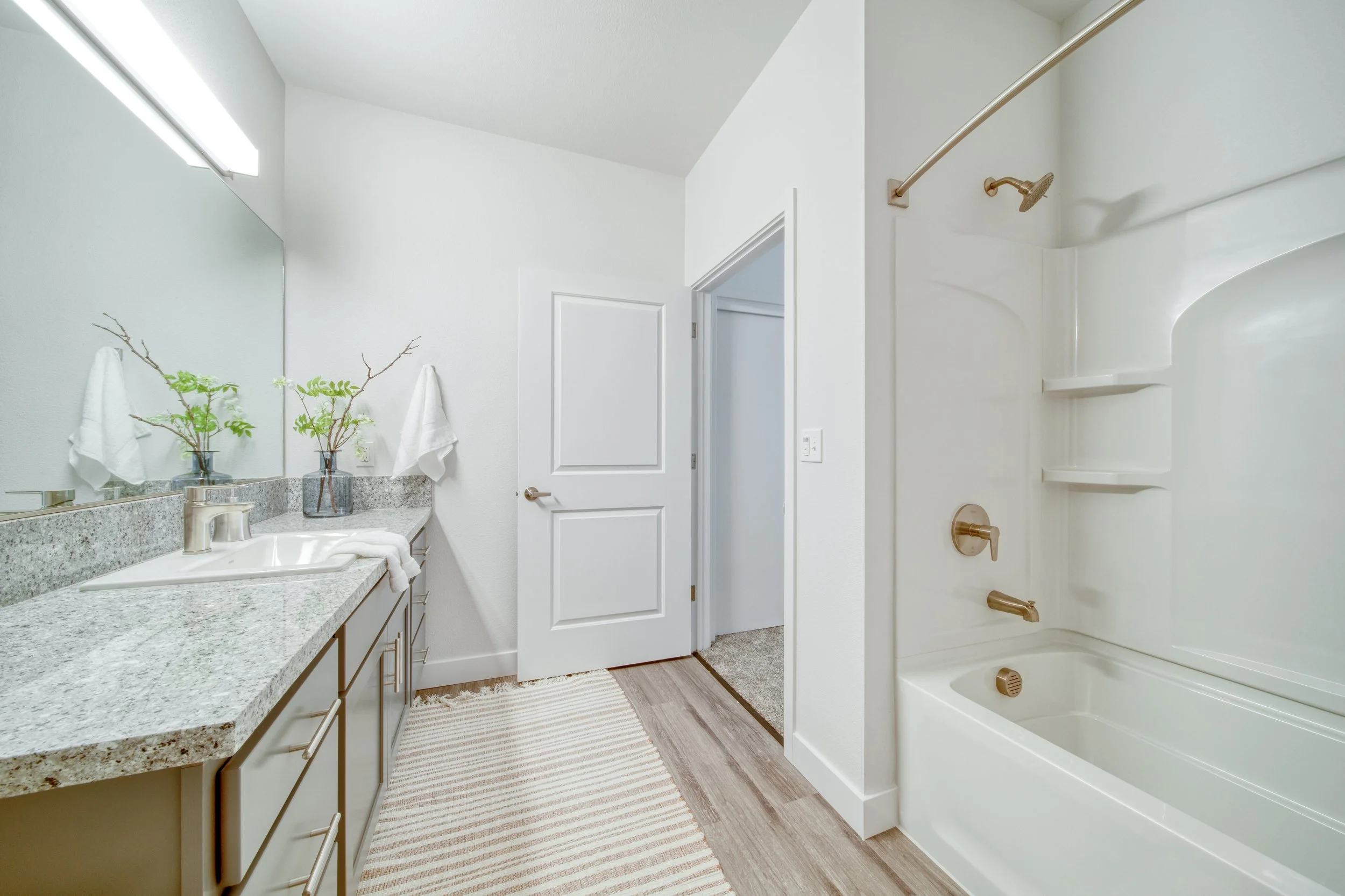 A modern bathroom with a granite countertop vanity, large mirror, white walls, and a shower/tub with a curved shower rod and built-in shelves.