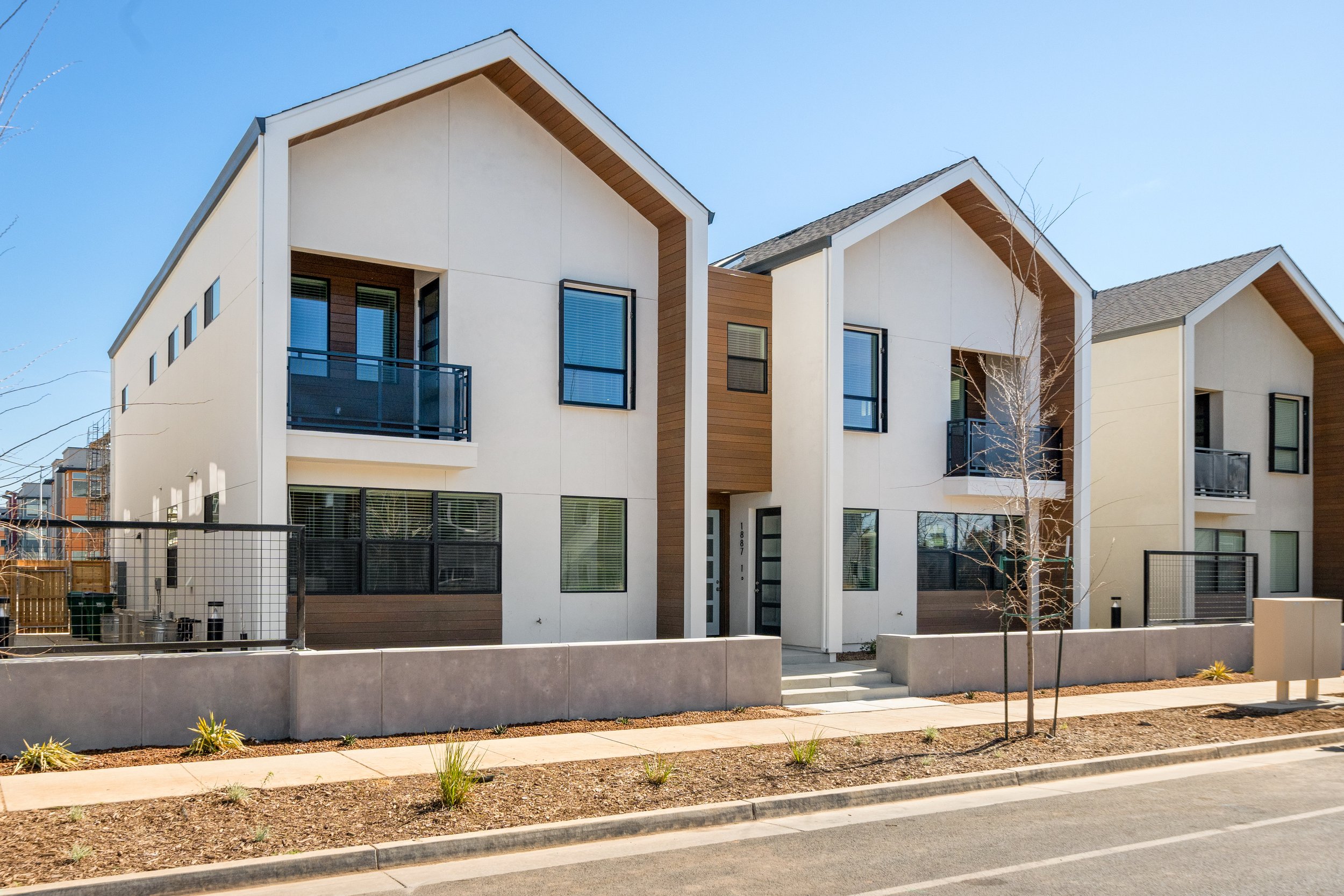 Modern multi-unit residential building with beige and brown siding, balconies, and large windows, situated along a sidewalk with small plants and a street in the foreground.