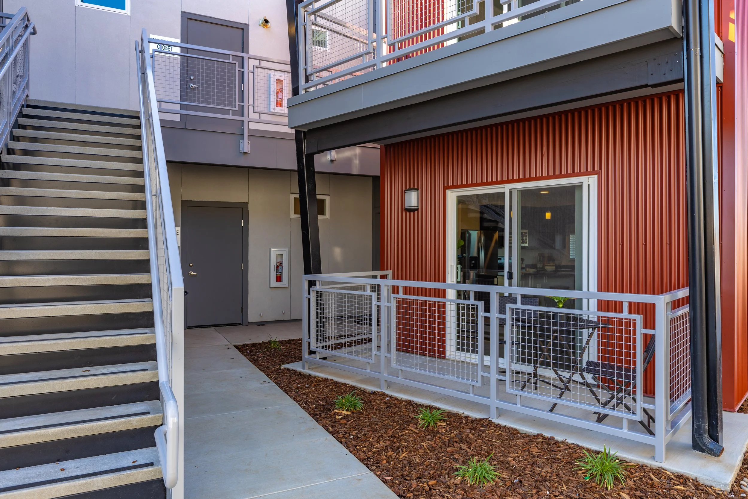 Exterior balcony area of a modern apartment building with grey metal railings, red corrugated wall, and a sliding glass door; adjacent to an outdoor staircase with a metal handrail leading to upper floors, with sidewalk and small landscaped garden wi