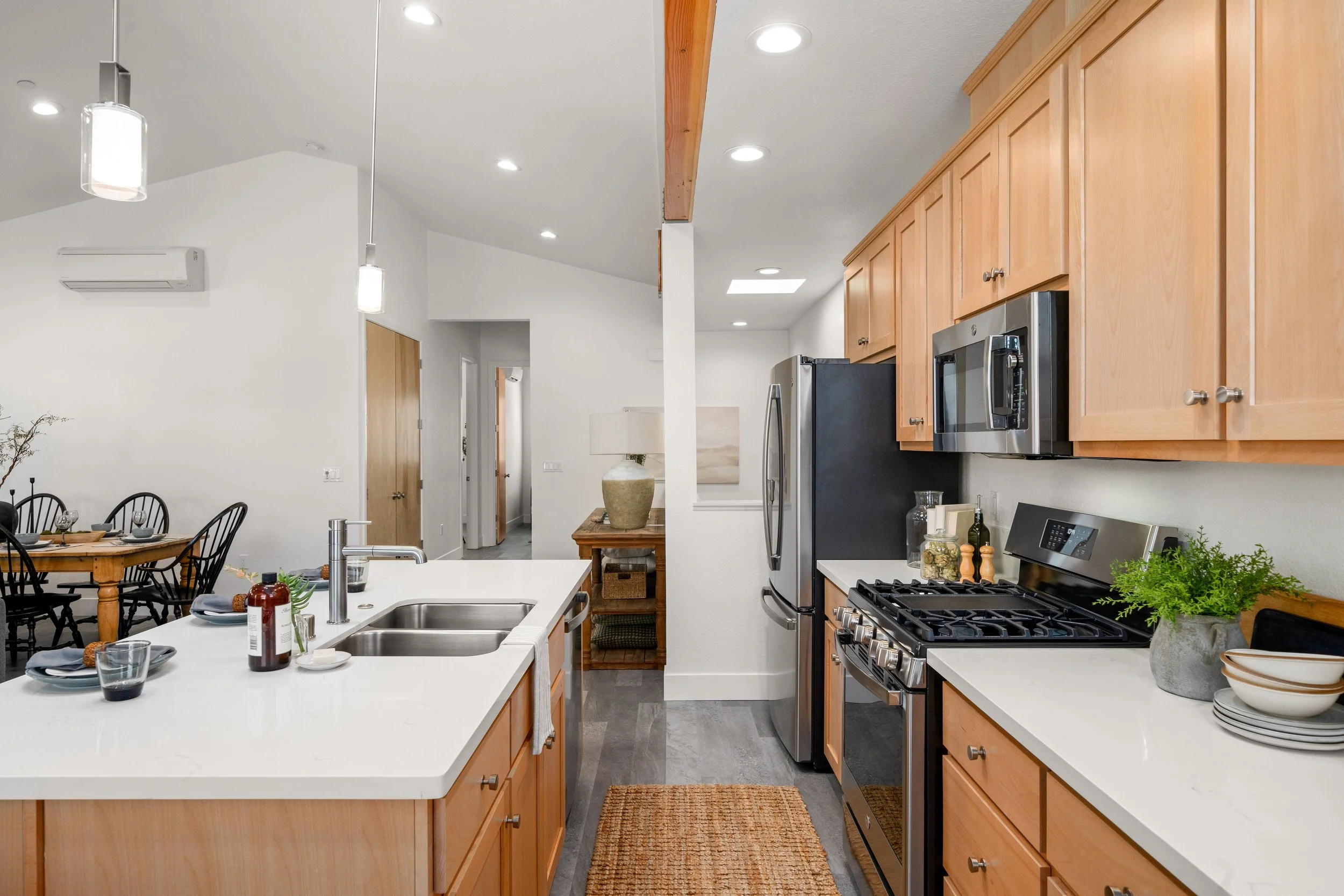Modern kitchen with light wood cabinets, stainless steel appliances including a refrigerator and microwave, white countertop island with sink and utensils, and a dining area with black chairs and wooden table in the background.