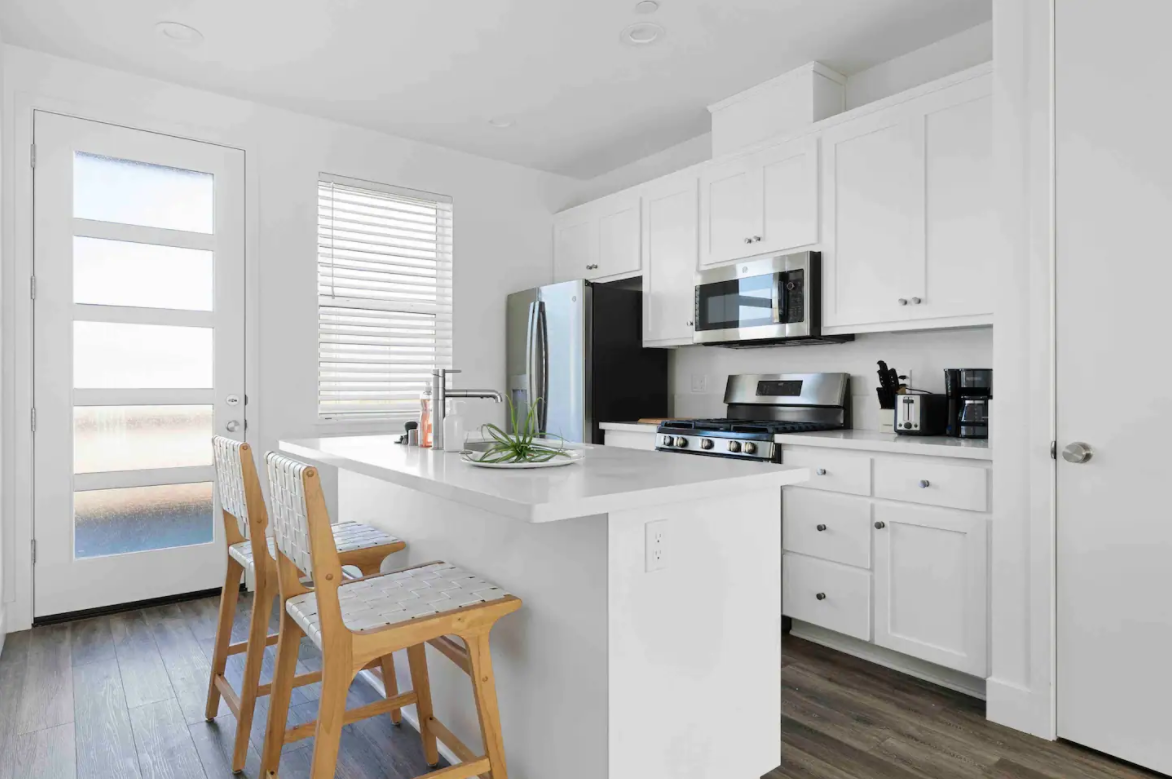 Modern kitchen with white cabinets, stainless steel appliances, a white kitchen island with barstools, and large windows allowing natural light.