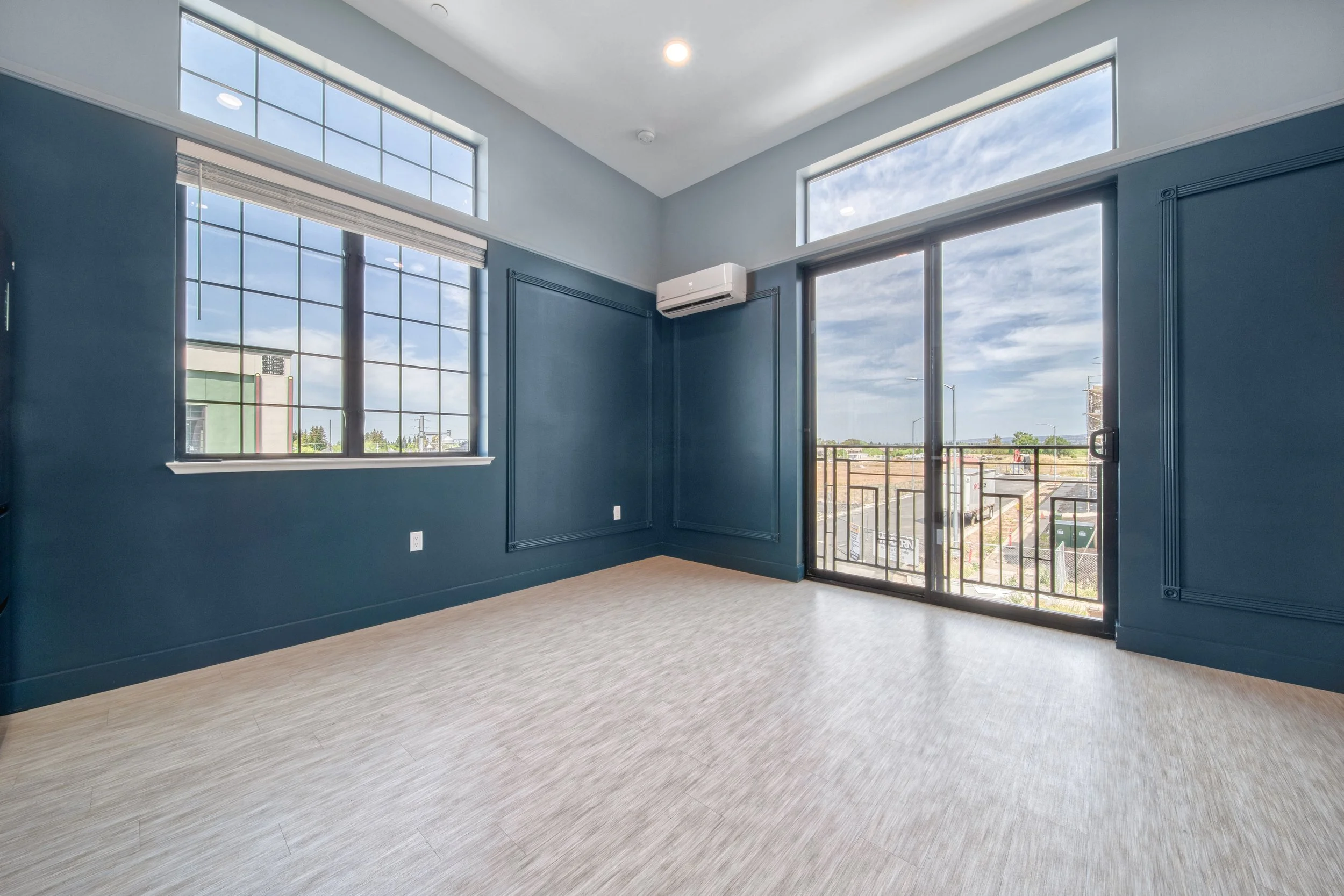 Empty room with blue walls, large windows, a sliding glass door leading to a balcony, and a view of a cityscape with blue sky and clouds.