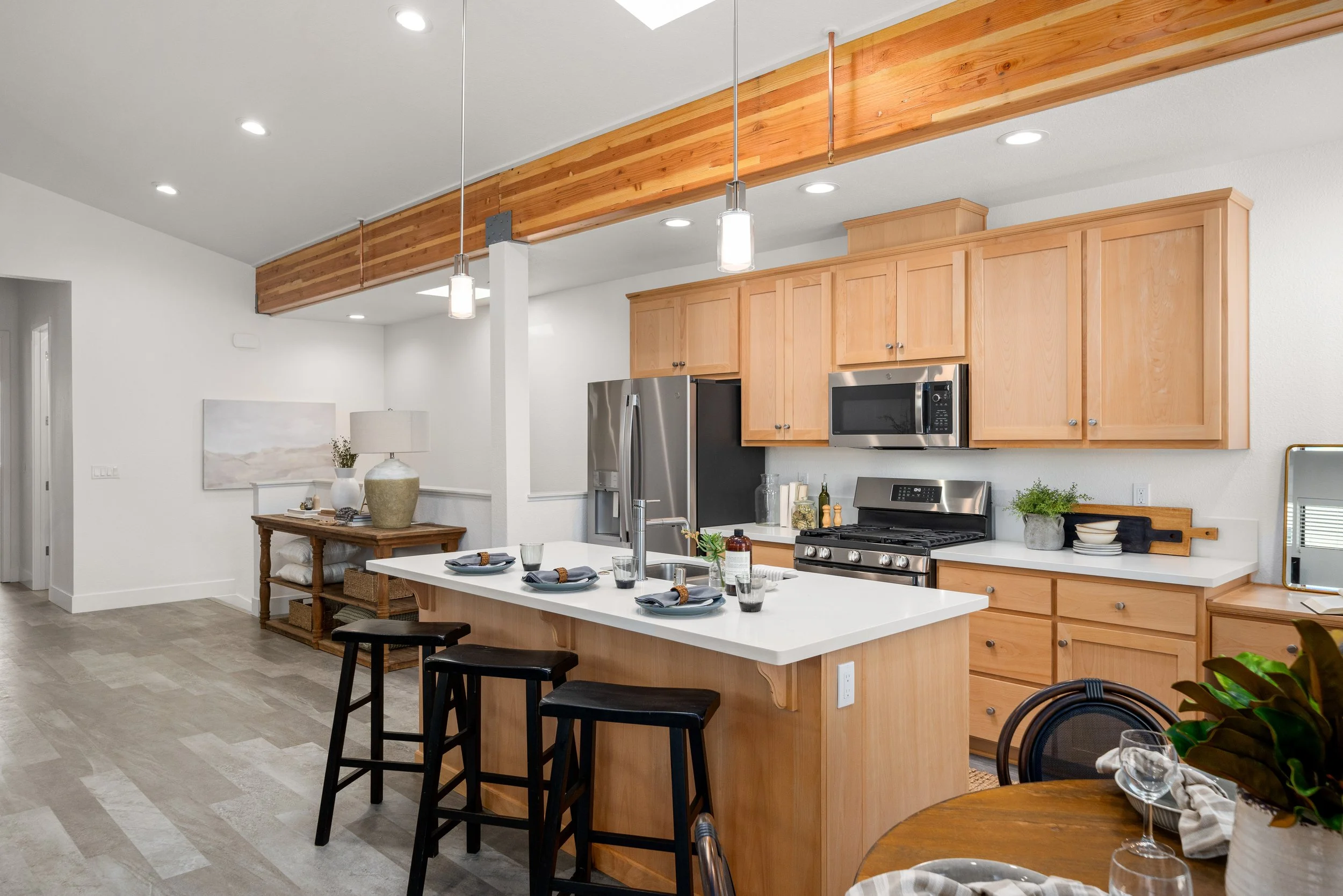Modern kitchen with light wood cabinets, stainless steel appliances, a white island with black stools, and a dining table set with glasses and a plant.