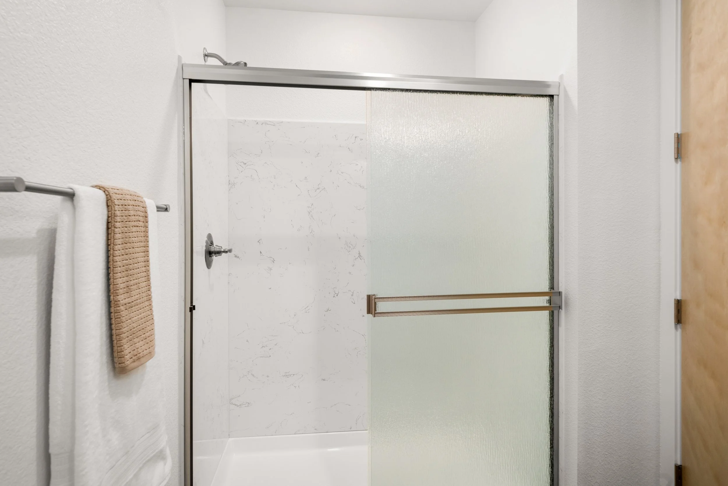 Bathroom shower with frosted glass sliding doors, beige towel on towel rack, white walls, wood door on right.