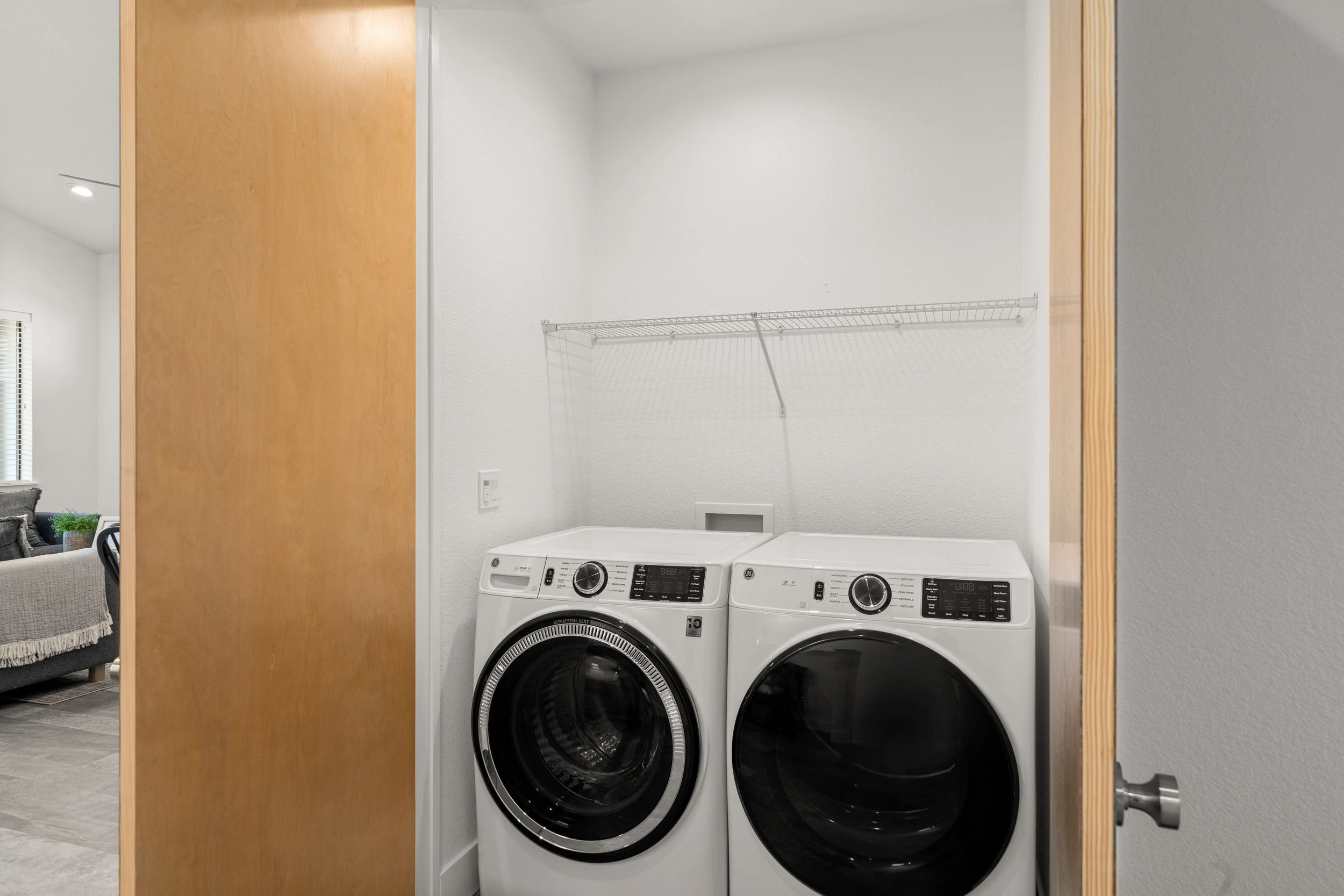 A laundry room with a washing machine and dryer side by side against a white wall, with a wire shelf above them, partially open wooden door on the left, and part of a living room with a sofa and window visible in the background.
