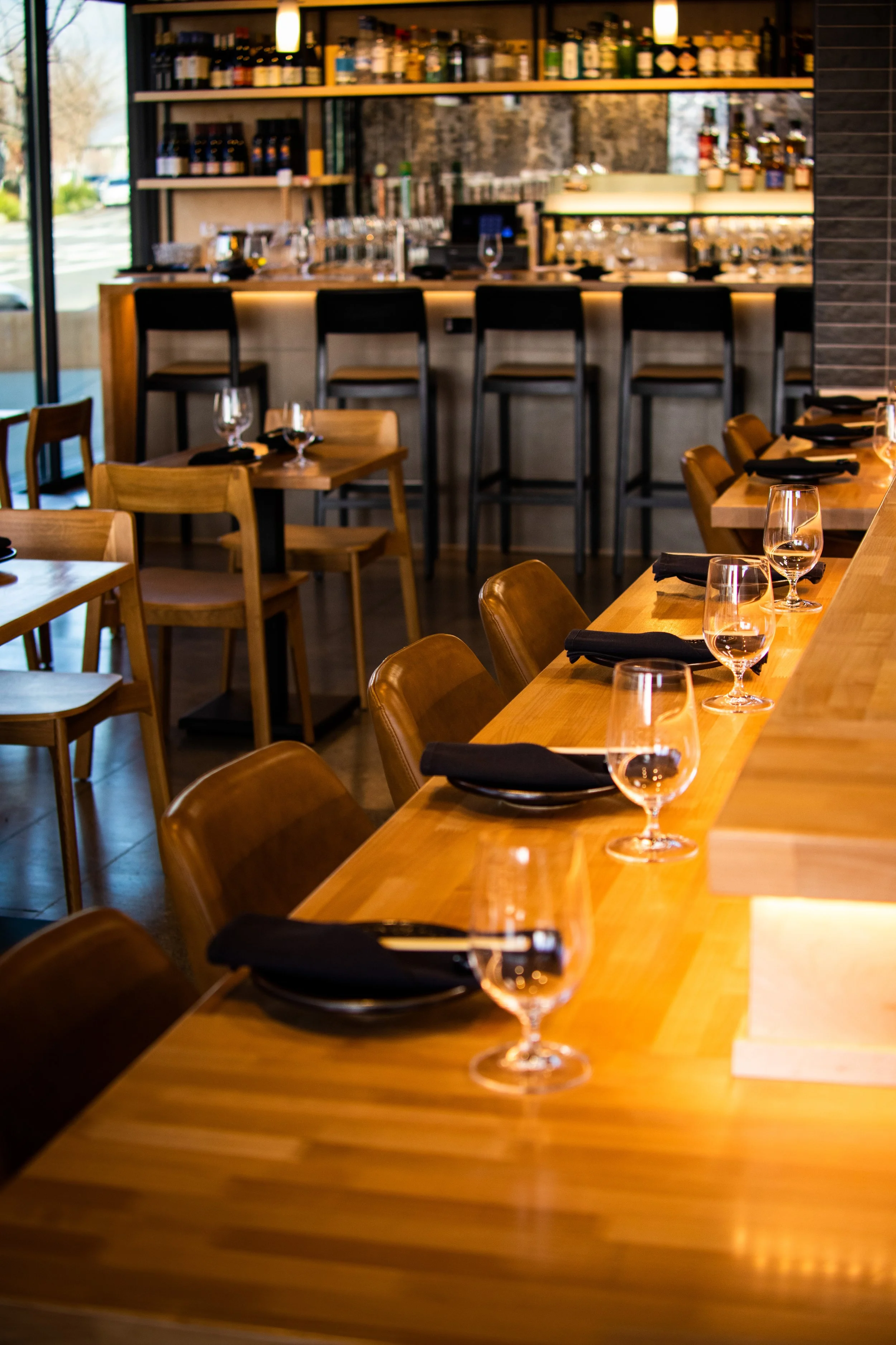 Empty restaurant dining area with wooden tables, black napkins, and wine glasses set at each place. There is a bar in the background with barstools and shelves with bottles.