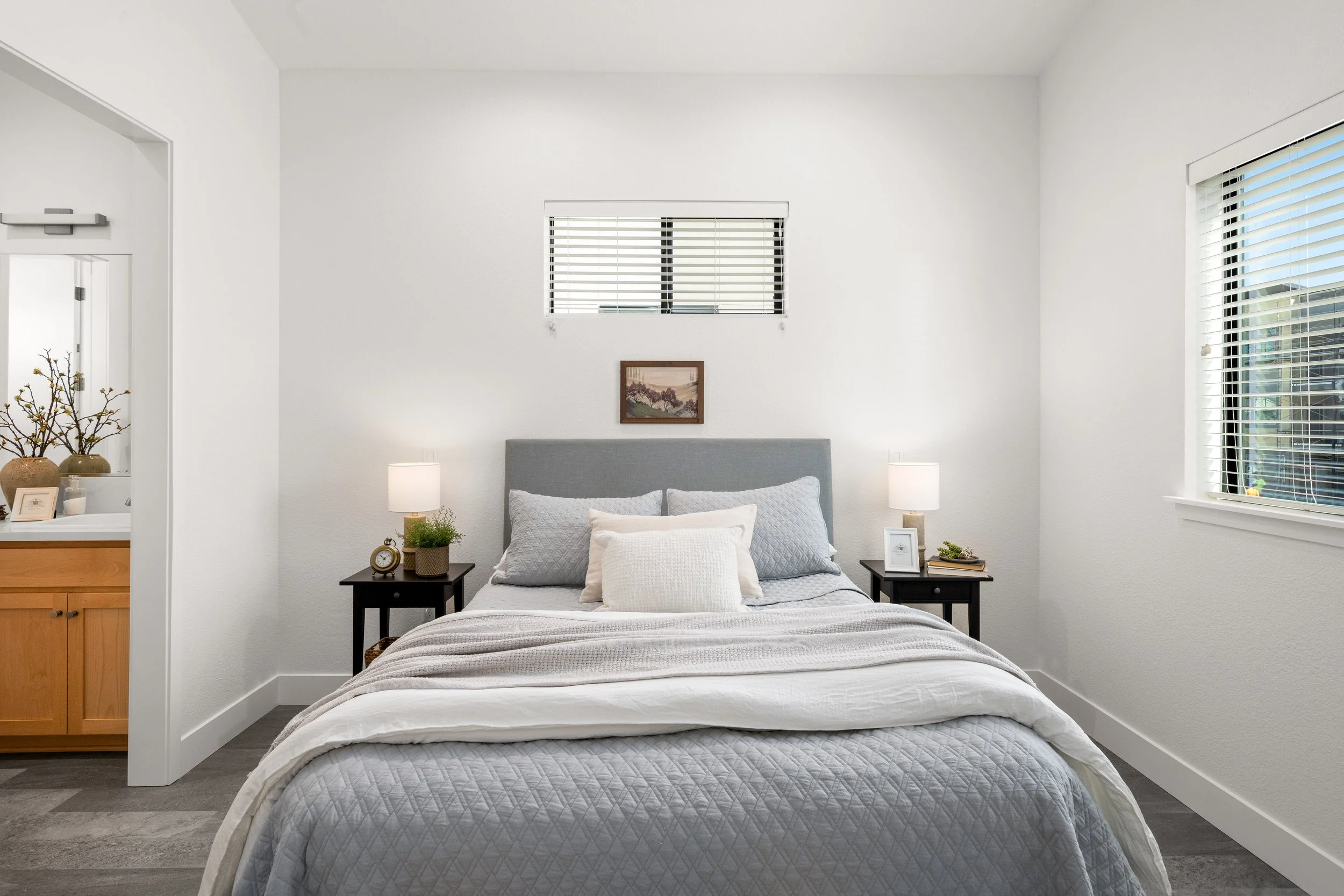 A neatly made bed with gray and white bedding, two pillows, and a headboard in a bright, minimally decorated bedroom with white walls, small windows with blinds, and two nightstands with lamps and decor.