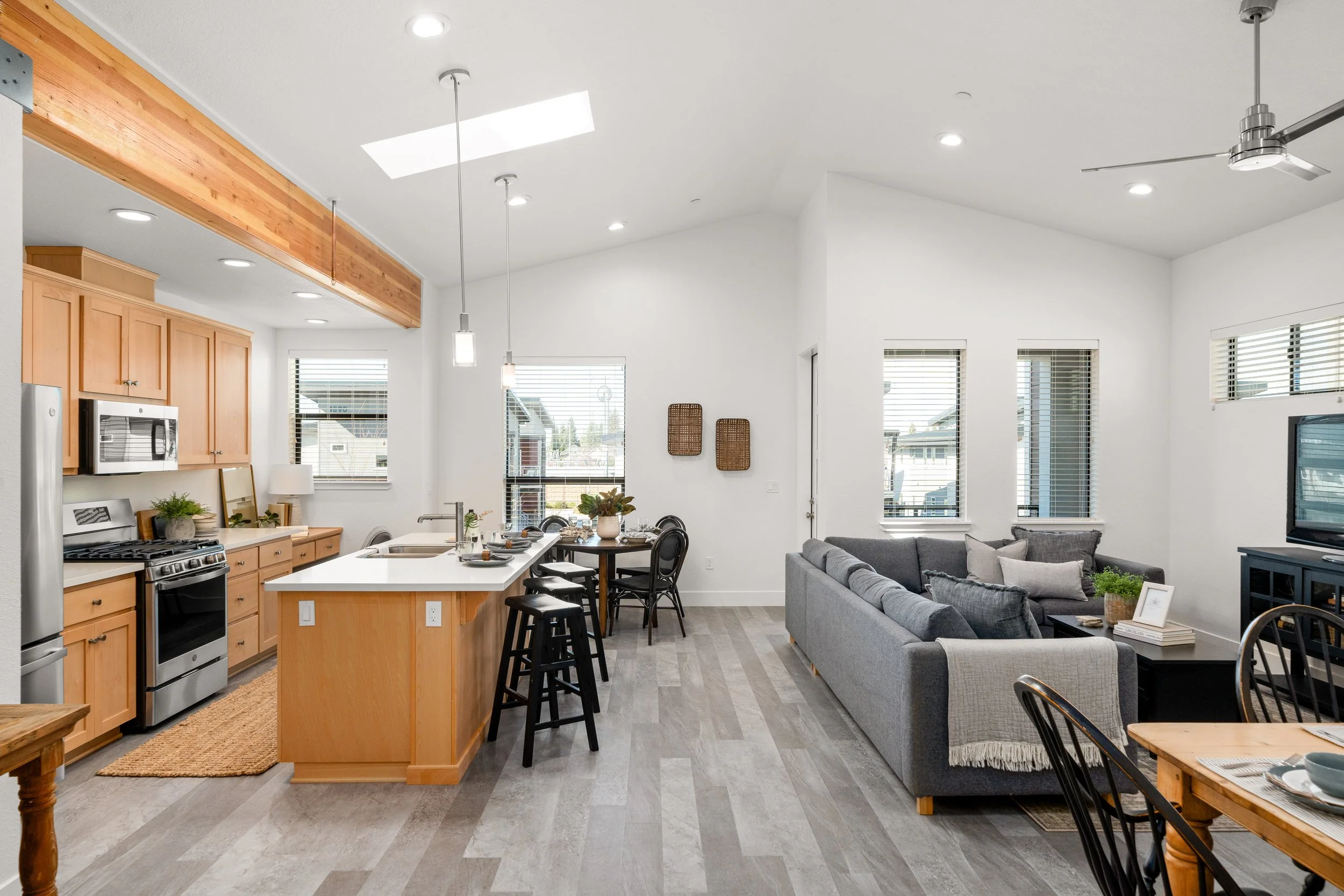 Open concept living room and kitchen with white walls, light wood cabinets, a gray sofa, and dining area with black chairs, natural light from multiple windows, and a ceiling fan.