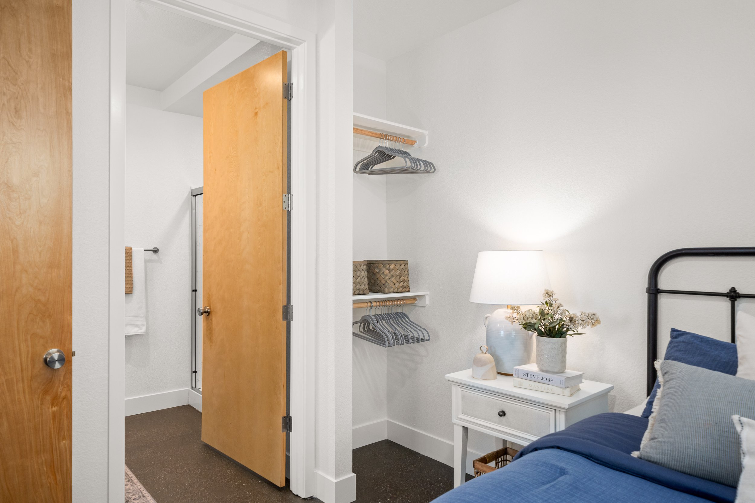 Bedroom with blue bed, white nightstand, lamp, decorative items, and open closet with hanging rods and baskets, adjacent to a bathroom with white walls and wooden door.