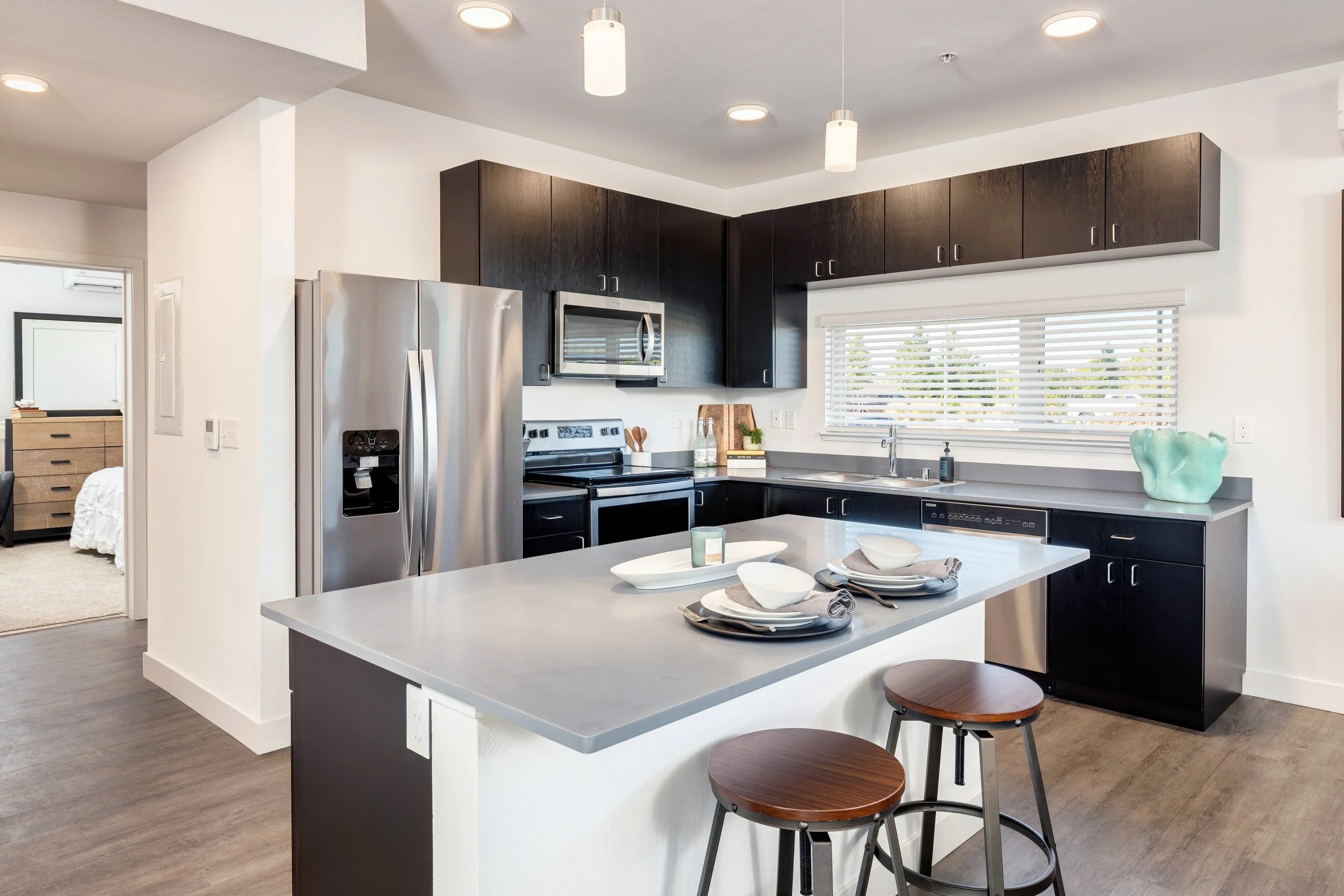 Modern kitchen with dark cabinets, stainless steel refrigerator, microwave, and dishwasher, white walls, large window with blinds, kitchen island with tableware, and two wooden stools.