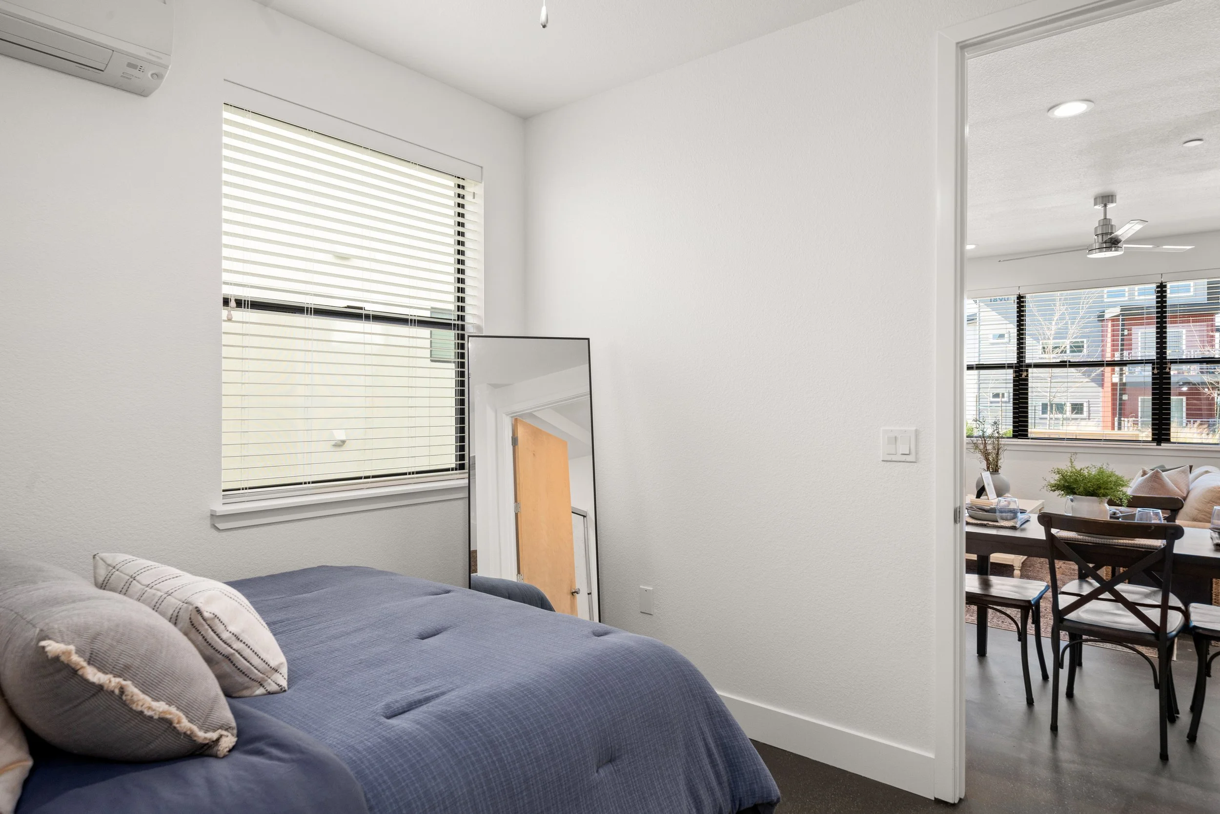 A bedroom with a bed covered in a blue quilt and pillows, a window with blinds, a leaning mirror, and an open doorway leading to a dining area with large windows, a table with chairs, and a ceiling fan.