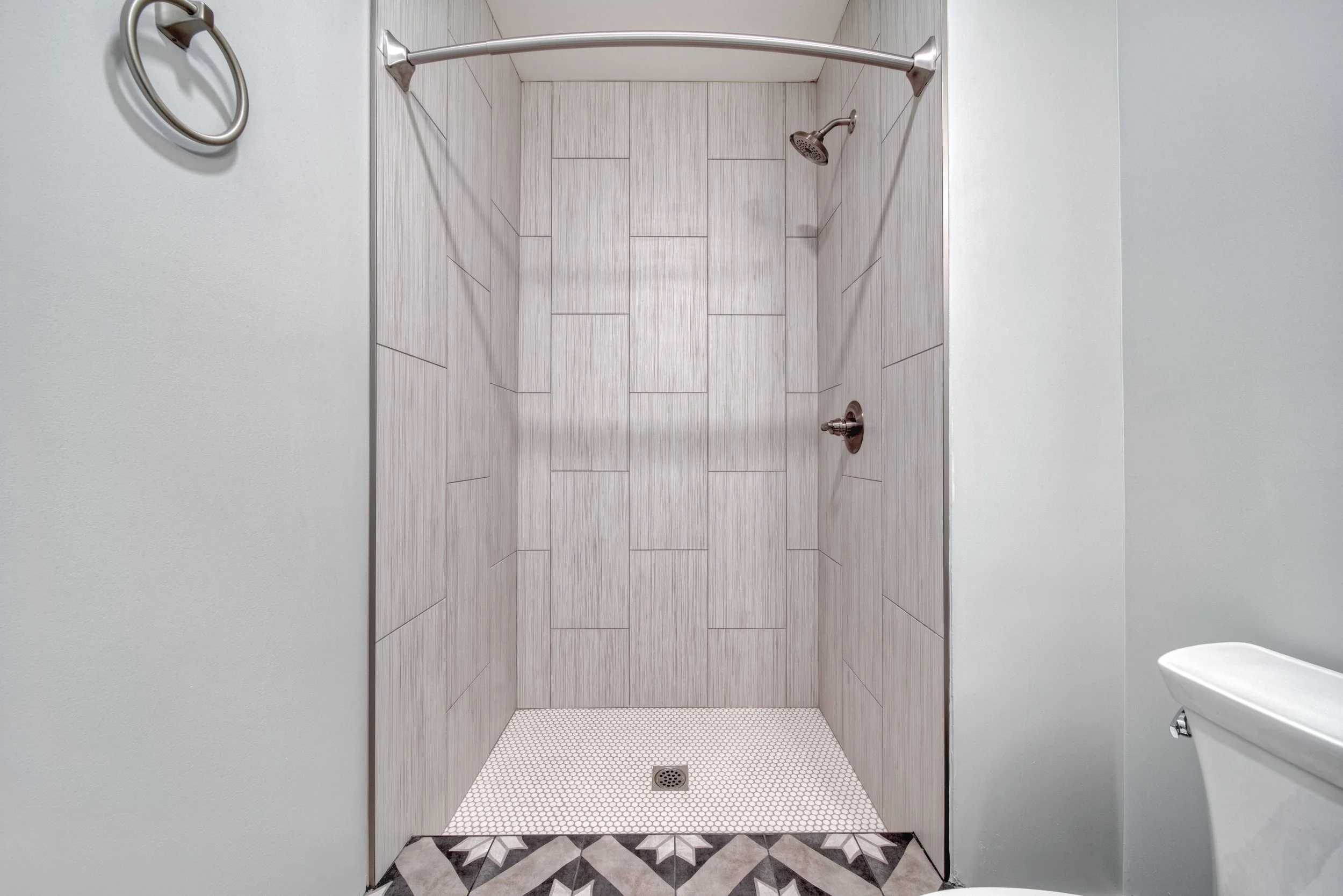 Empty shower with beige tiles, handheld shower head, shower faucet, metal towel ring on white wall, and partial view of toilet at the right.