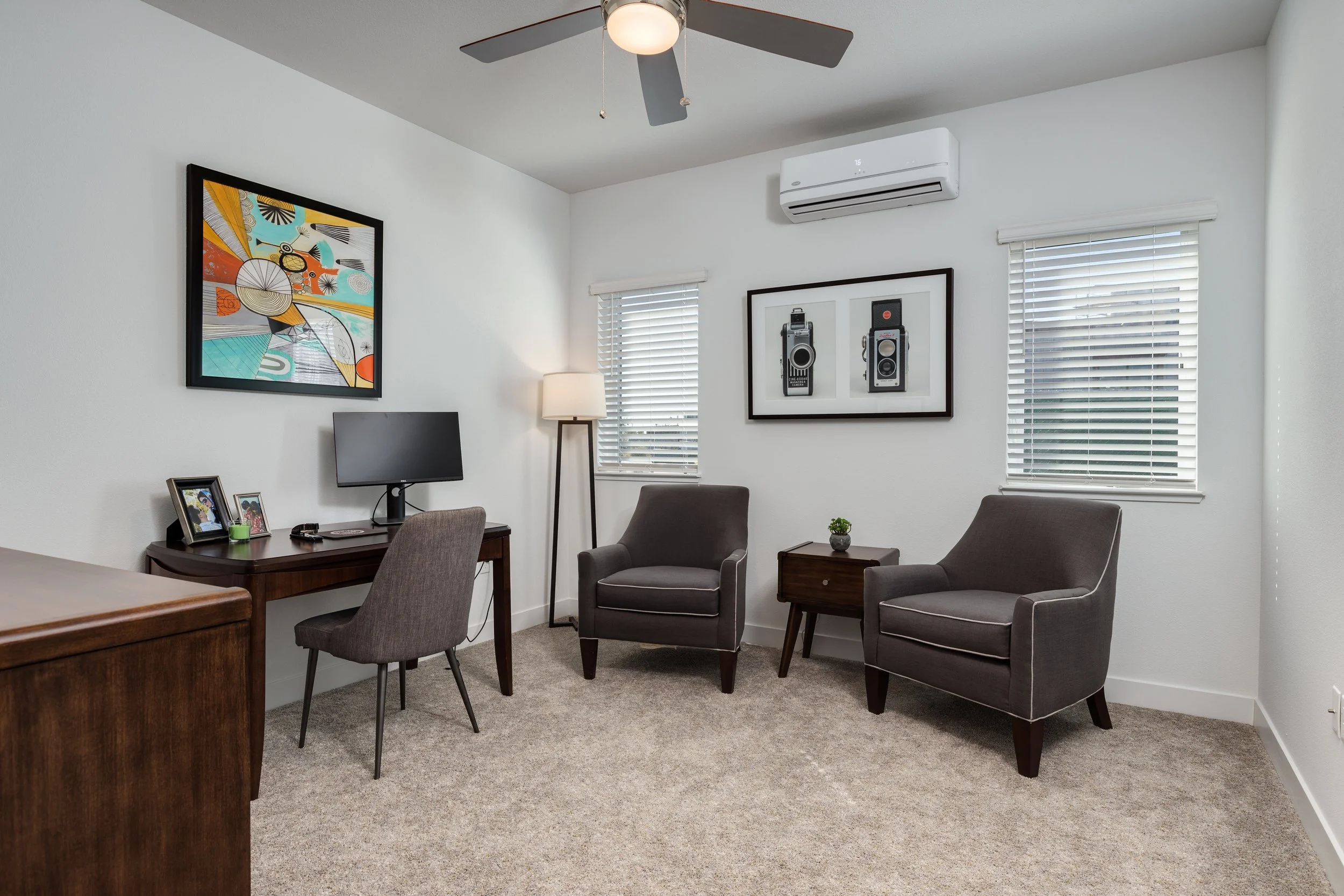 A home office with two gray armchairs, a wooden side table, and a small plant. There is a desk with a computer monitor, framed photos, and a lamp. The room has two windows with white blinds, artwork on the walls, a ceiling fan, and a white air condit