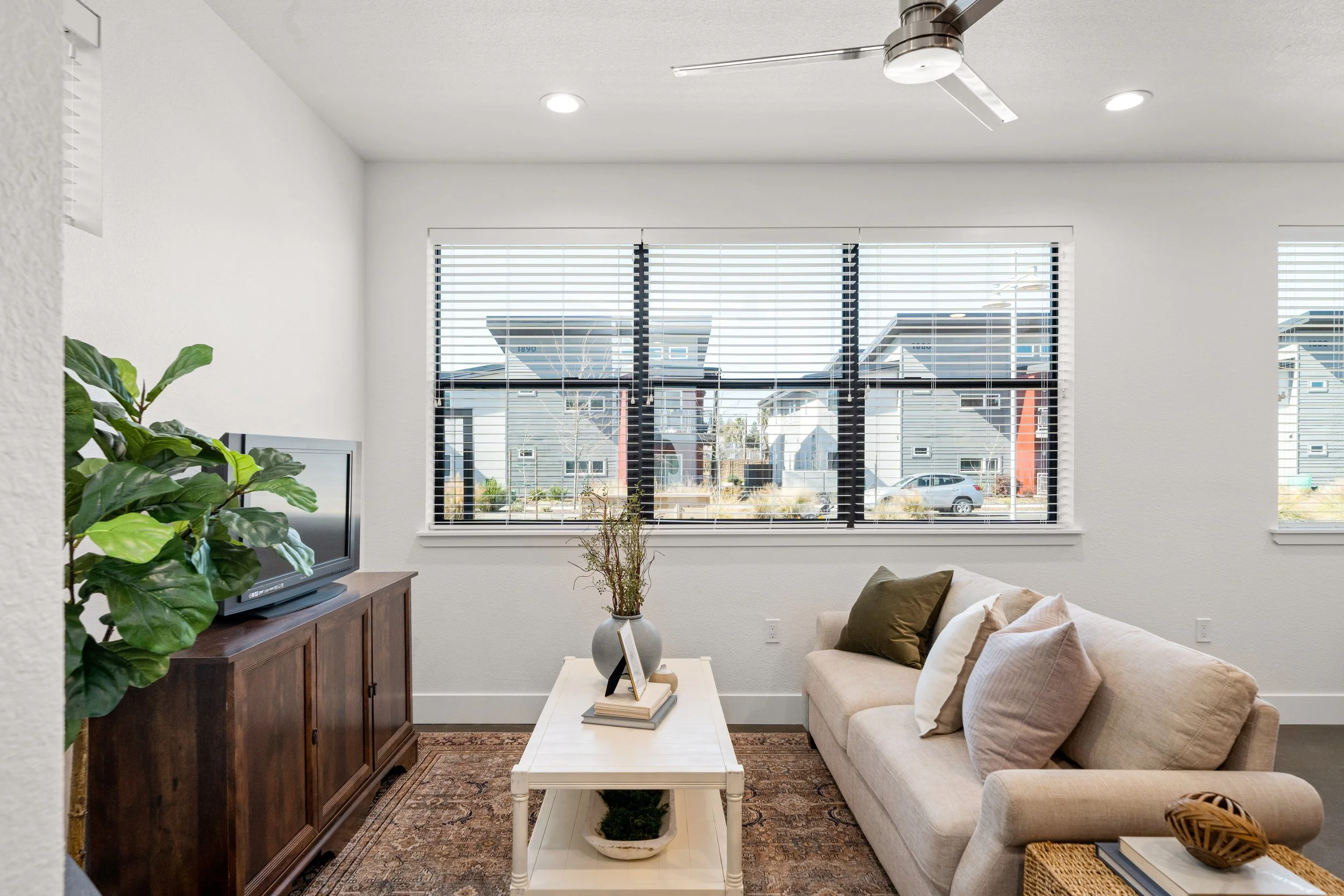 A cozy living room with a beige sofa, decorative pillows, a white coffee table with books and a plant, a dark wooden media console with a TV, and large windows with blinds showing a neighborhood outside. There is a ceiling fan and recessed lighting.
