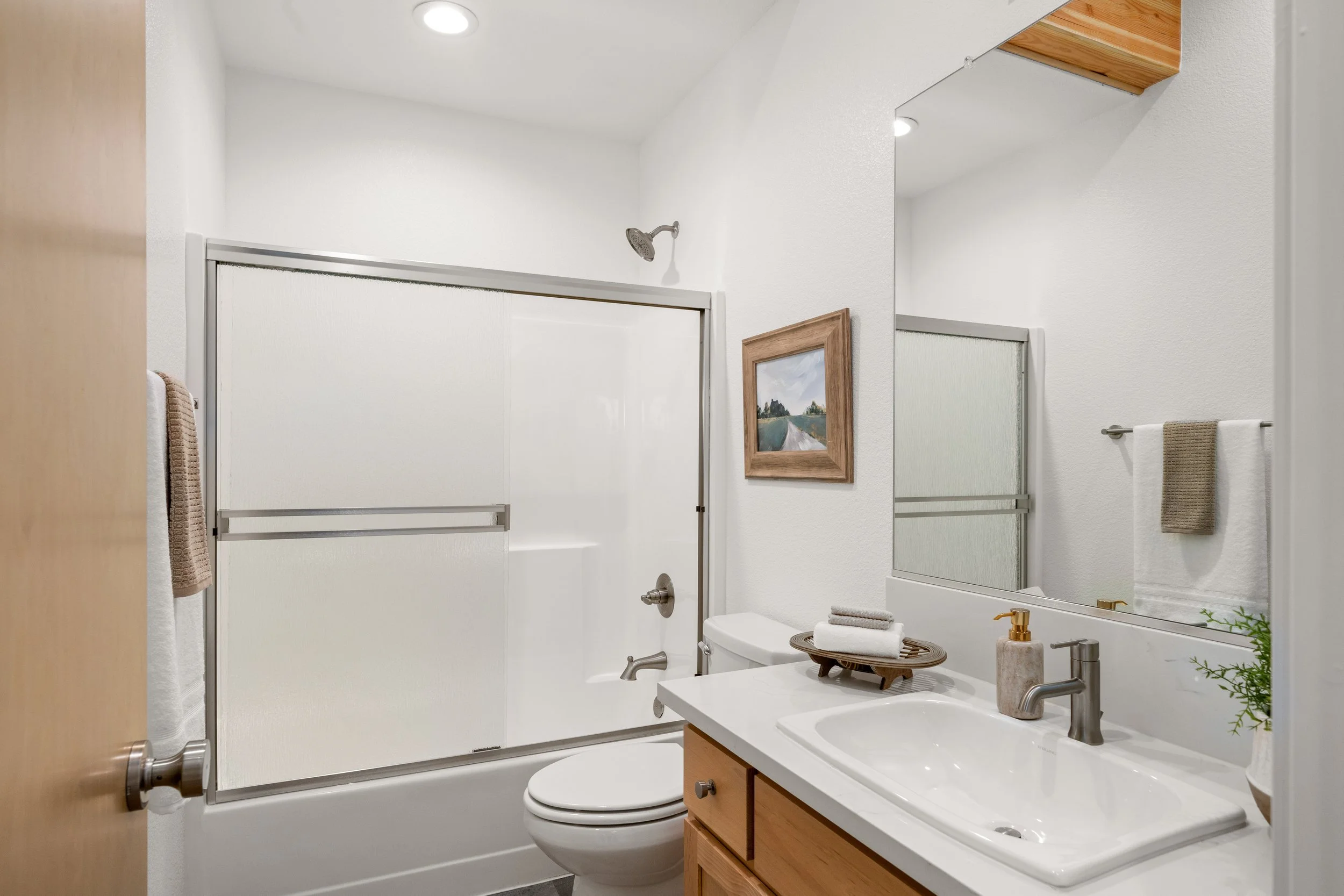Clean bathroom with white walls, a bathtub with a sliding door, a toilet, a wooden vanity with a sink, a large mirror, towels on a rack, a framed painting on the wall, and a small plant.