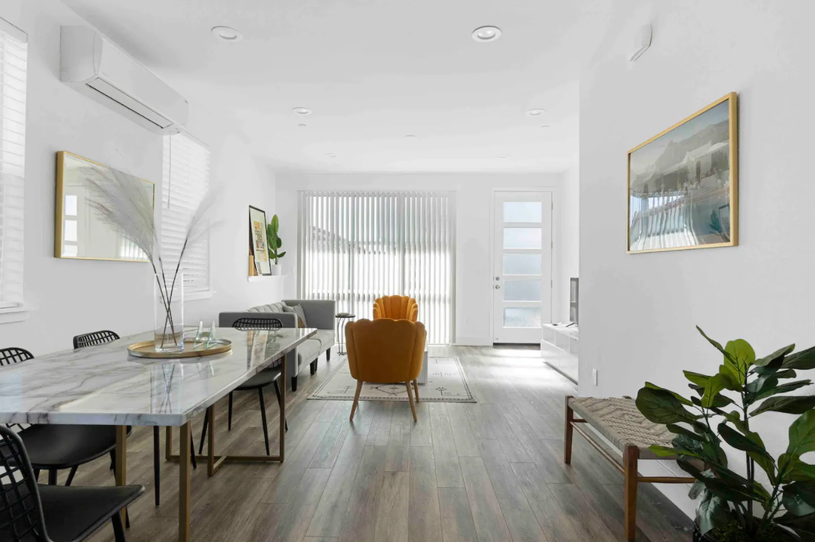 Bright living room with white walls, wooden flooring, a marble dining table, black chairs, a gray sofa, an orange armchair, a large window with vertical blinds, art on the walls, and indoor plants.