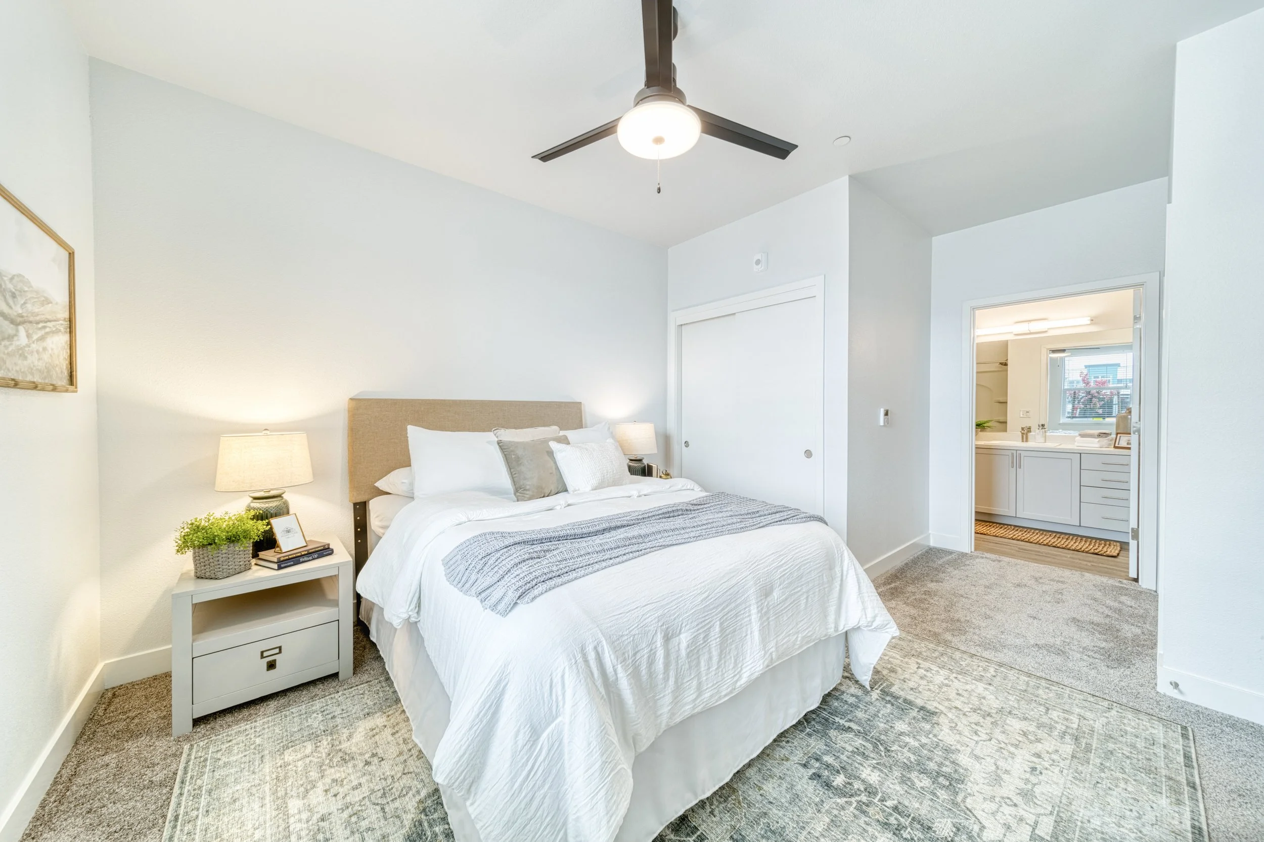 A bedroom with a white bed, multiple pillows, a beige headboard, and white bedding. There are two nightstands with lamps on each side of the bed, one with a plant and books. The room has a ceiling fan, a large rug, and a doorway leading to a bathroom