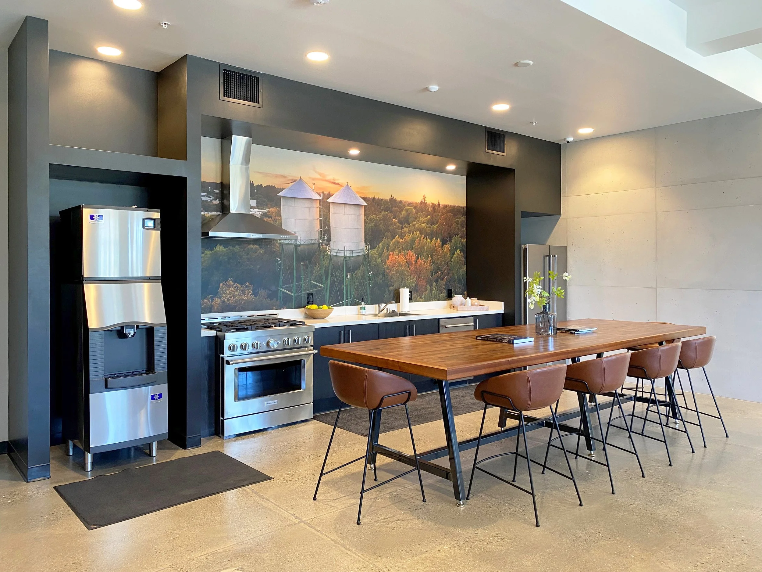 Modern kitchen with a black and stainless steel refrigerator, a metallic range hood, a stainless steel stove, black cabinets, and a large wooden dining table with brown chairs. A mural of silos and a sunset with trees is on the back wall.