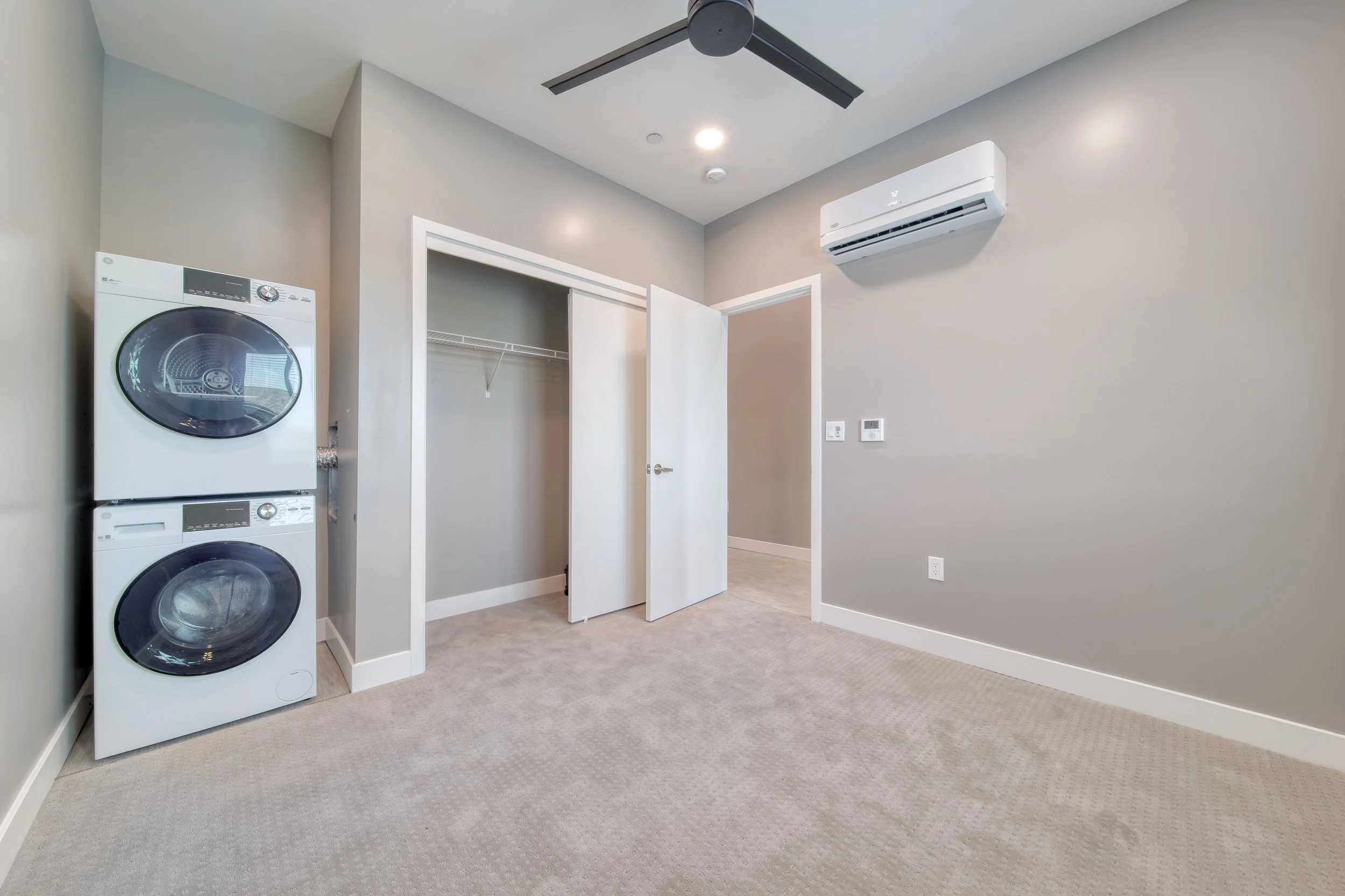 Empty laundry room with stacked washer and dryer, open closet, gray walls, ceiling fan, and wall-mounted air conditioner.