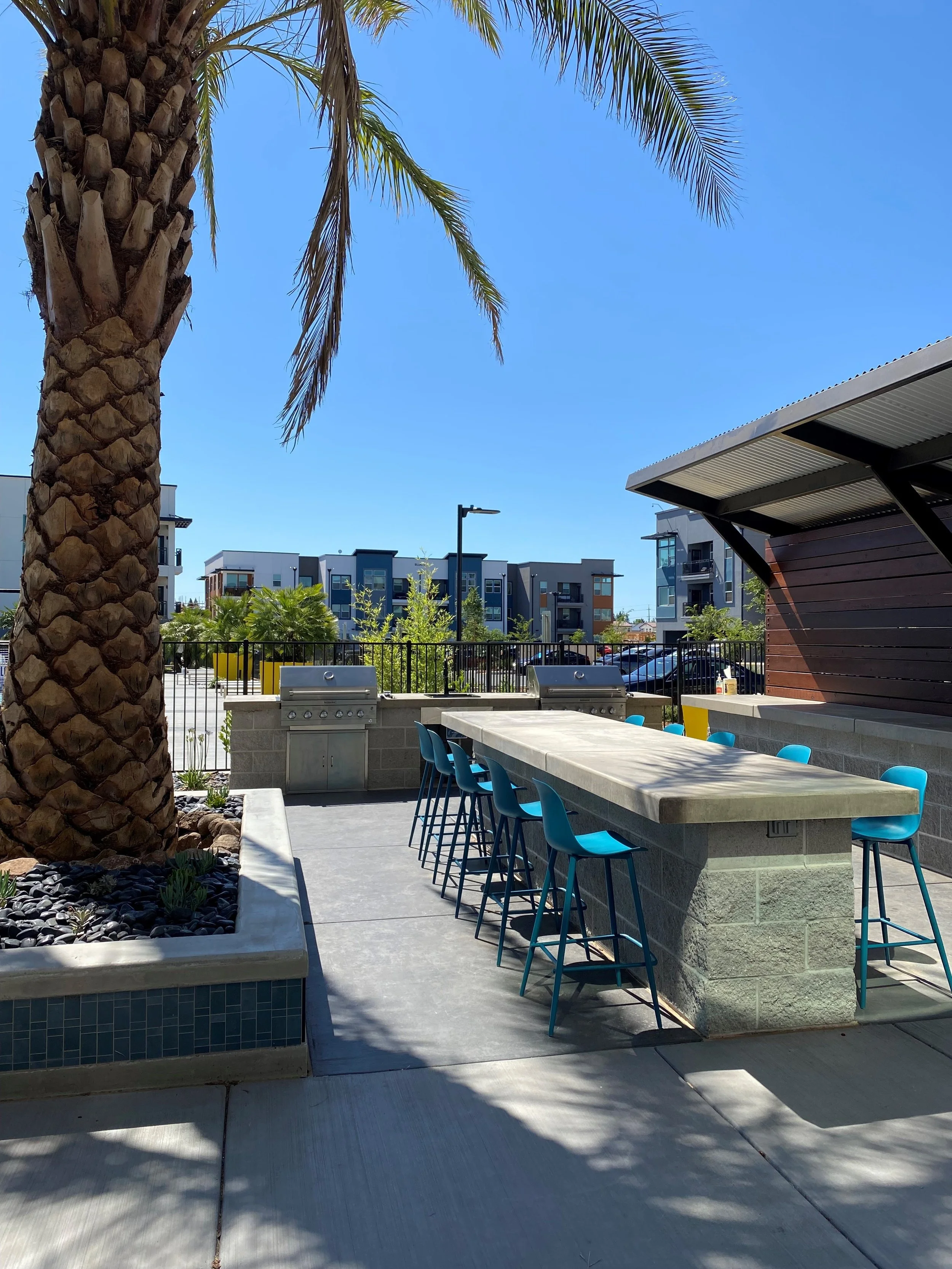 Outdoor poolside bar area with a long concrete bar counter, blue bar stools, and palm trees, in front of modern residential buildings.
