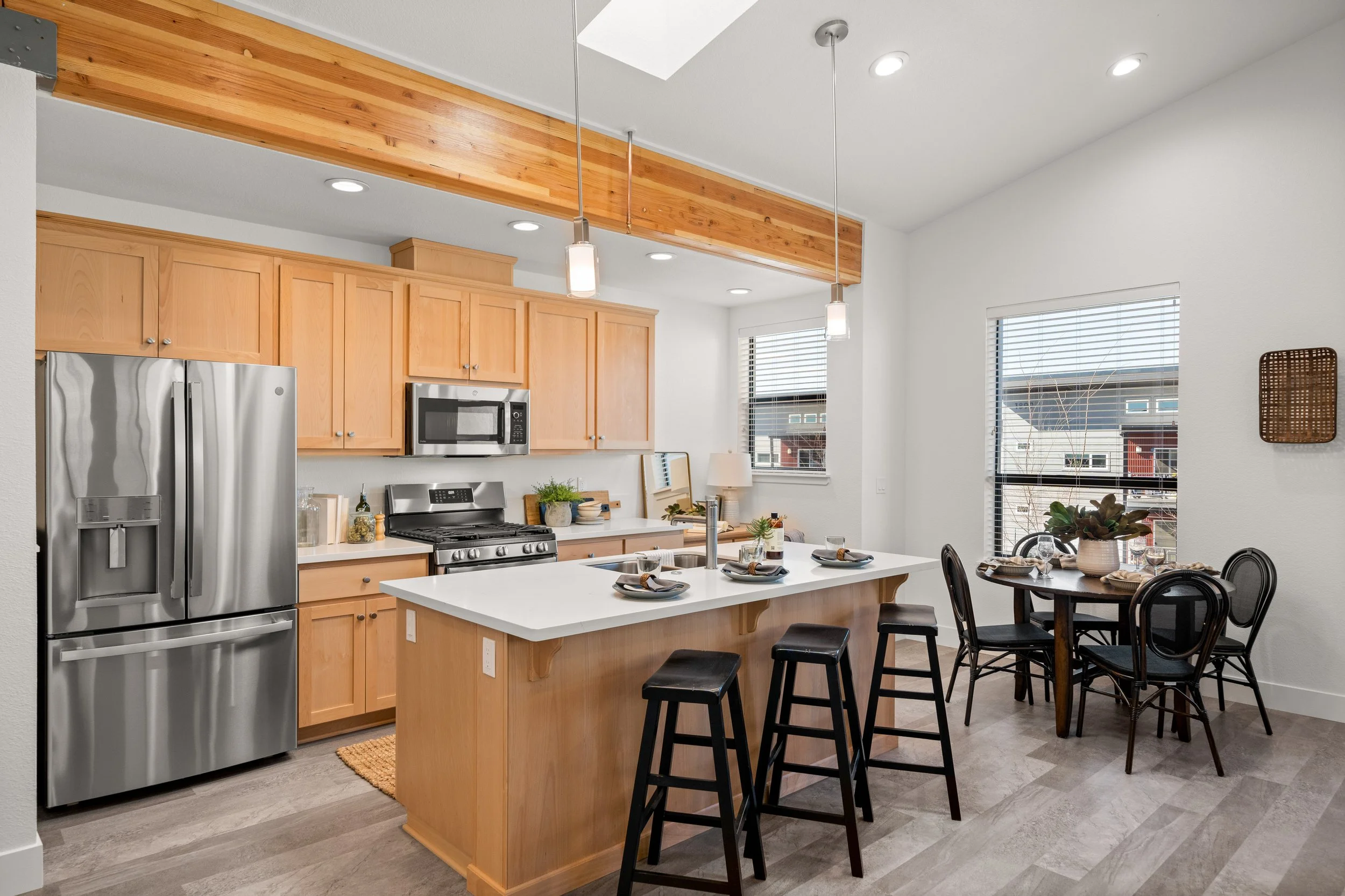 Kitchen with light wood cabinets, stainless steel refrigerator, microwave, and stove; white island with kitchenware, black stools; round dining table with black chairs, potted plant, and windows with blinds.