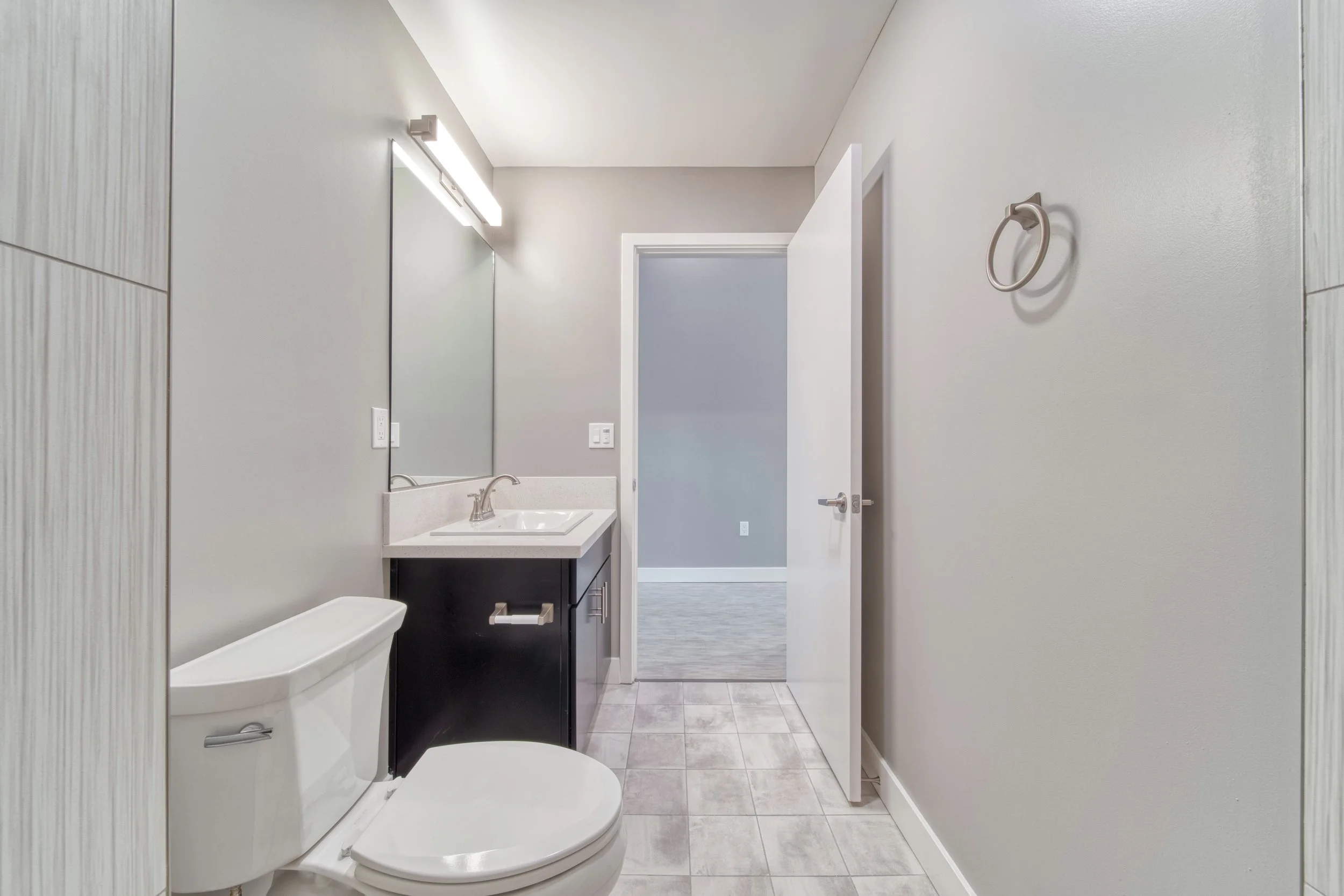 A modern bathroom with white walls and a tiled floor, featuring a toilet, a vanity with a sink, a mirror, and a towel ring on the wall.