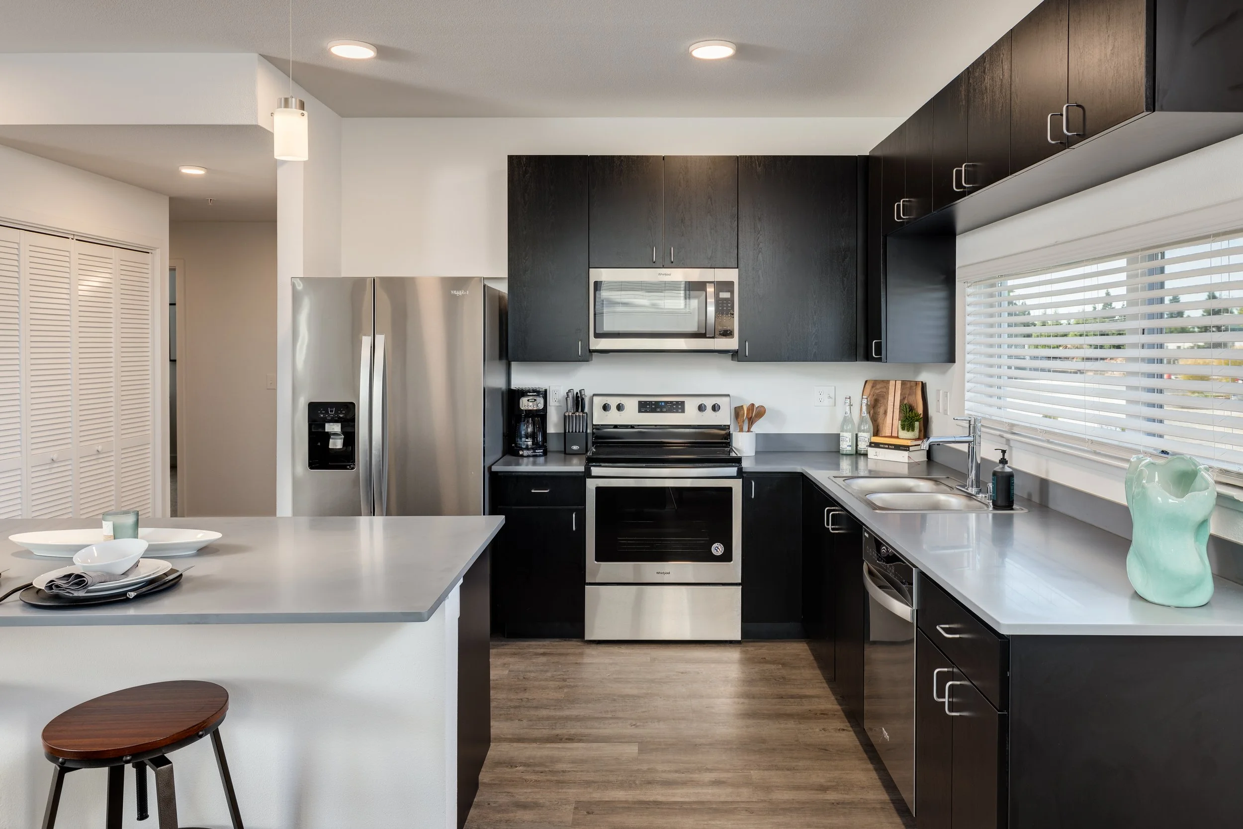Modern kitchen with black cabinets, stainless steel appliances including refrigerator, oven, microwave, and dishwasher. It features a large window with blinds and a kitchen island with a stool.