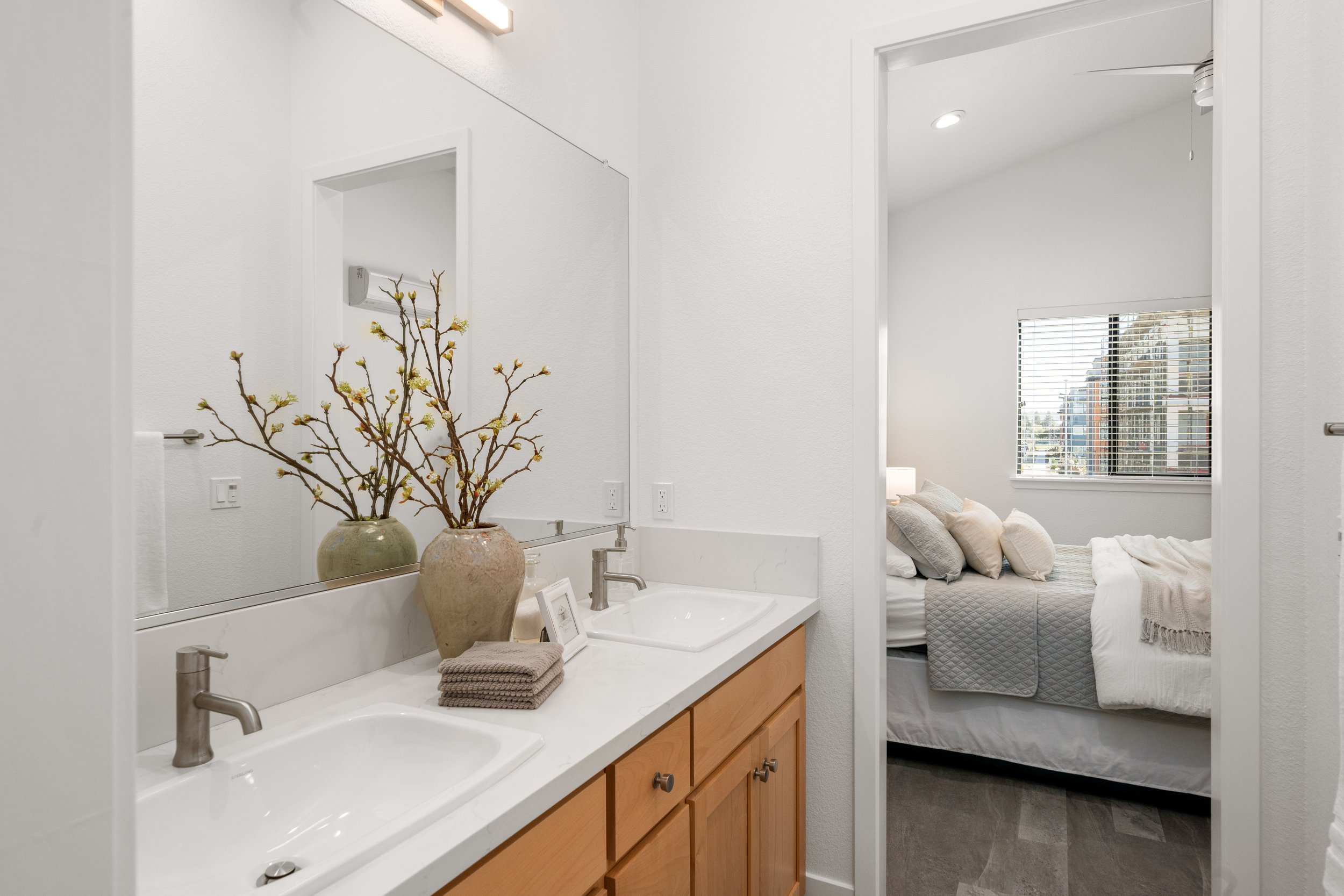Bathroom with a double sink vanity, large mirror, decorative vases with branches, and towels; view into a bedroom with a bed, pillows, and a window with blinds.