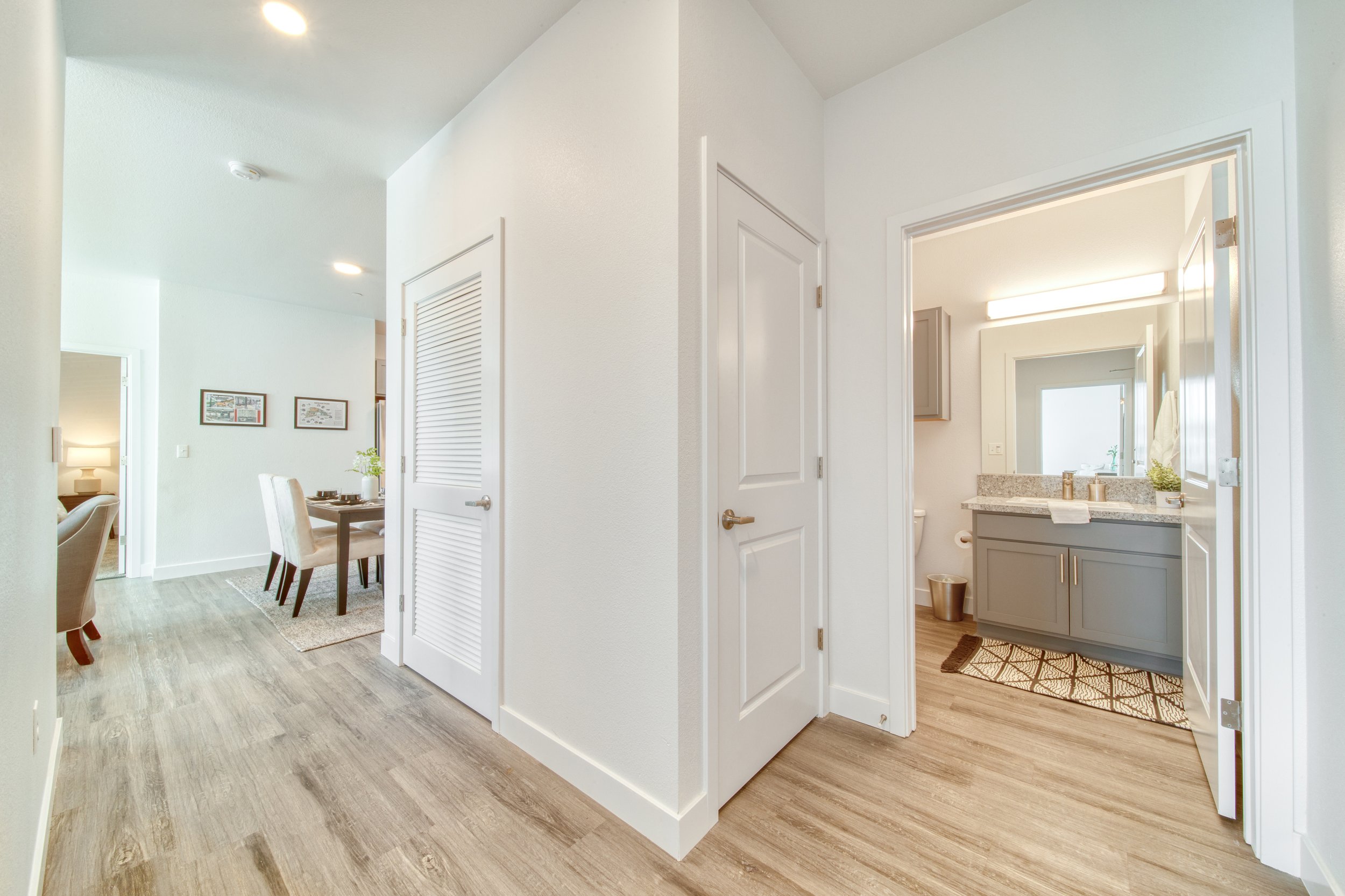 Interior view of a modern apartment with a hallway leading to a bathroom with gray cabinets and granite countertop, a mirrored mirror, and a window. A dining area with a white chair and a table is visible in the background.