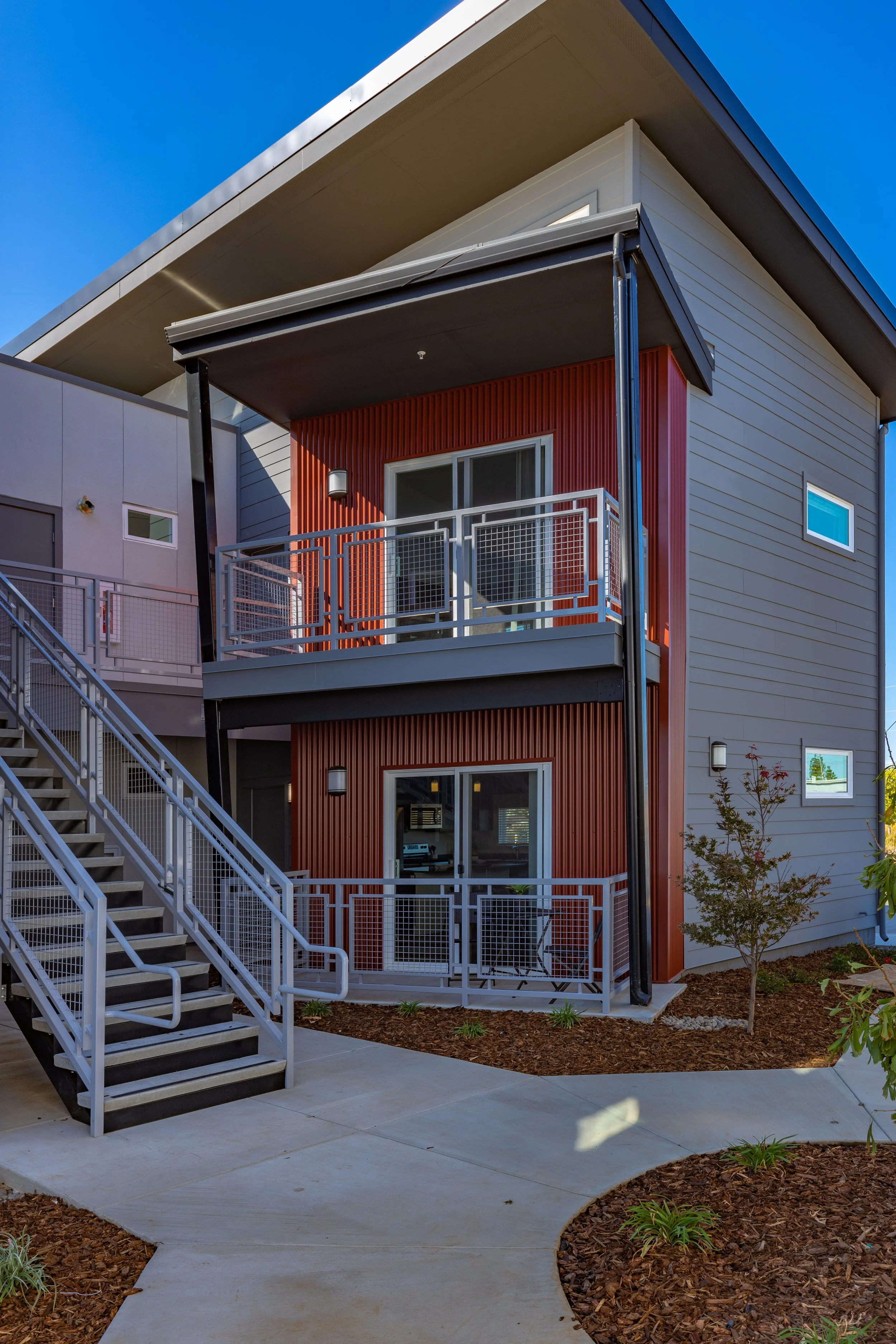 Exterior view of a modern, multi-story apartment building with gray and red siding, outdoor balconies, and an outdoor staircase, set against a clear blue sky.