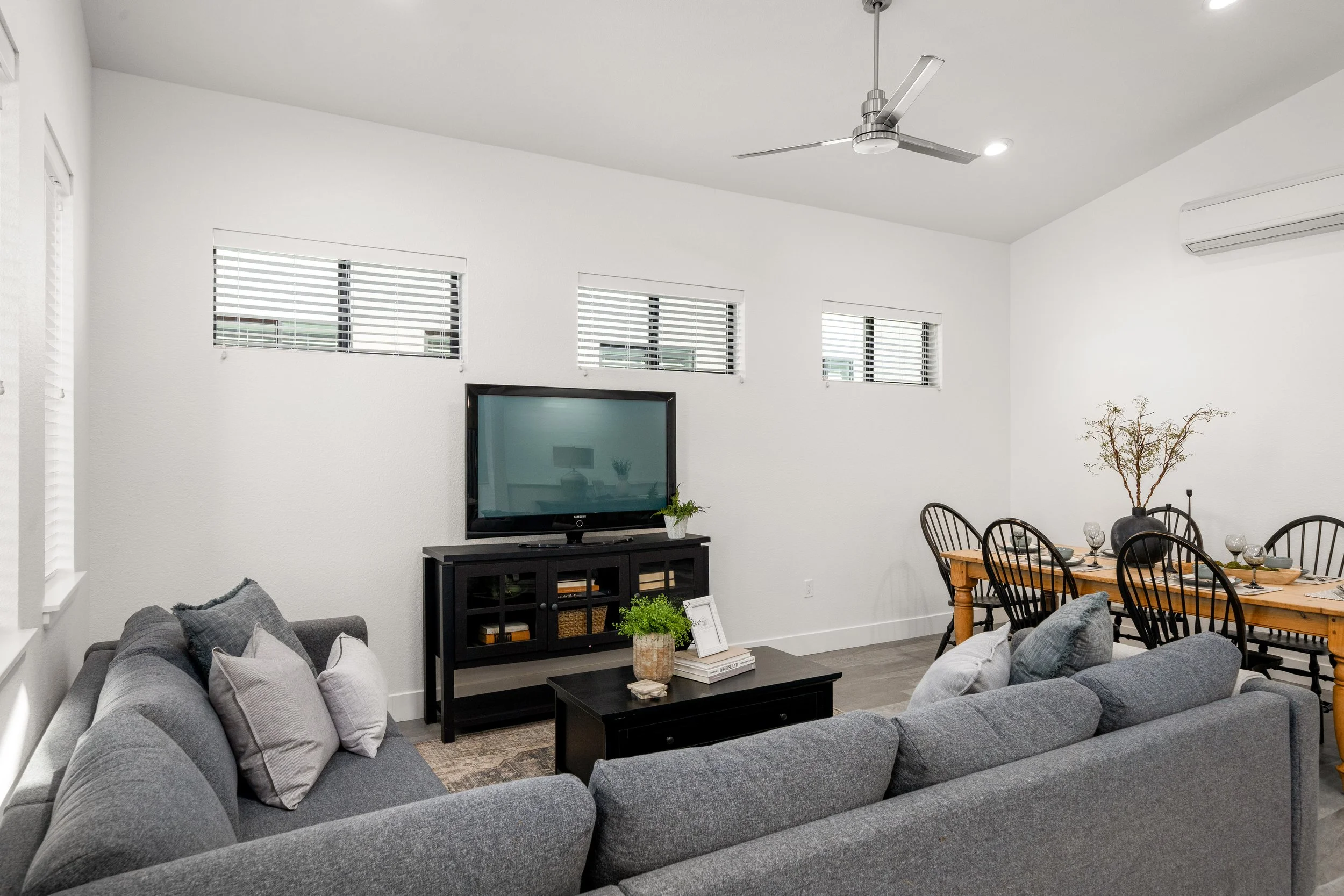 Living room with gray sectional sofa, black TV stand with a television, dining table with black chairs, ceiling fan, air conditioning unit, and small windows with blinds.