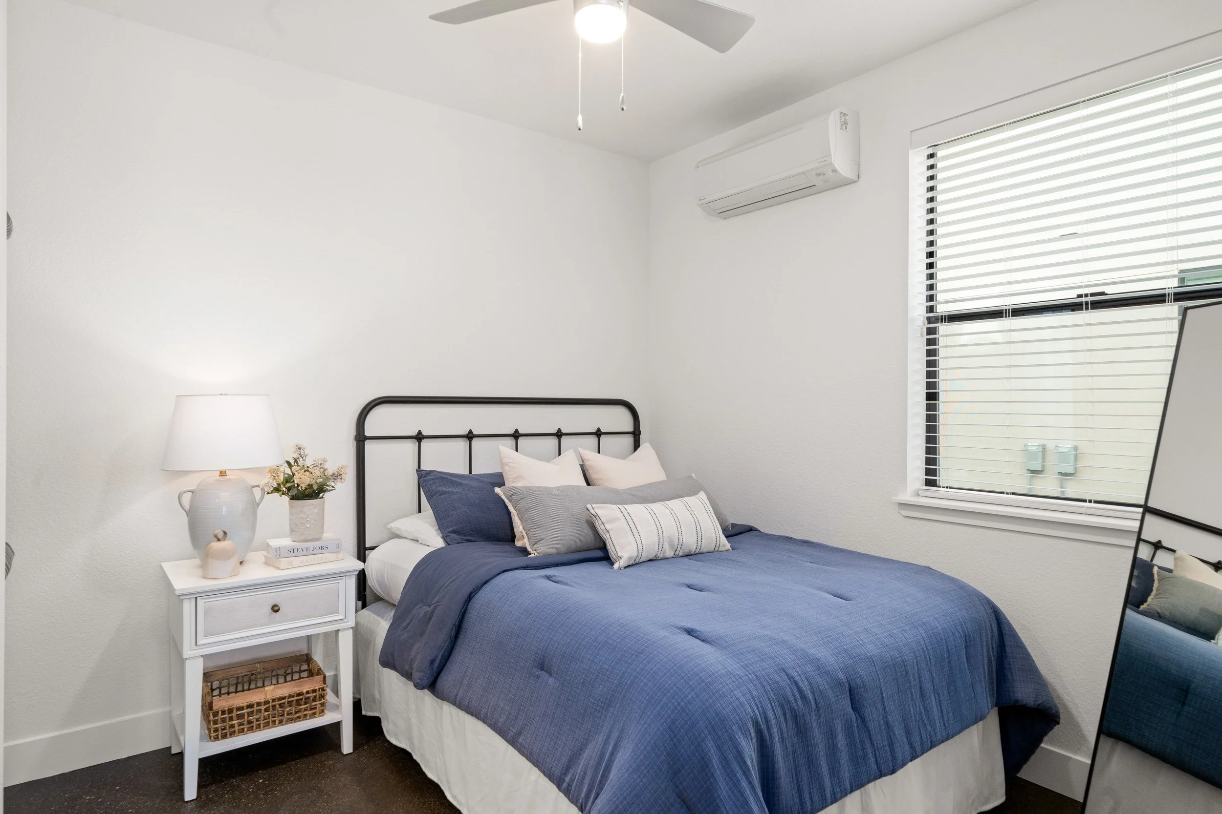 A neatly made bedroom with a queen-sized bed, blue bedding, and multiple pillows. Beside the bed is a white nightstand with a lamp, a small plant, and books. There is a window with blinds and an air conditioning unit above. A floor mirror is partiall