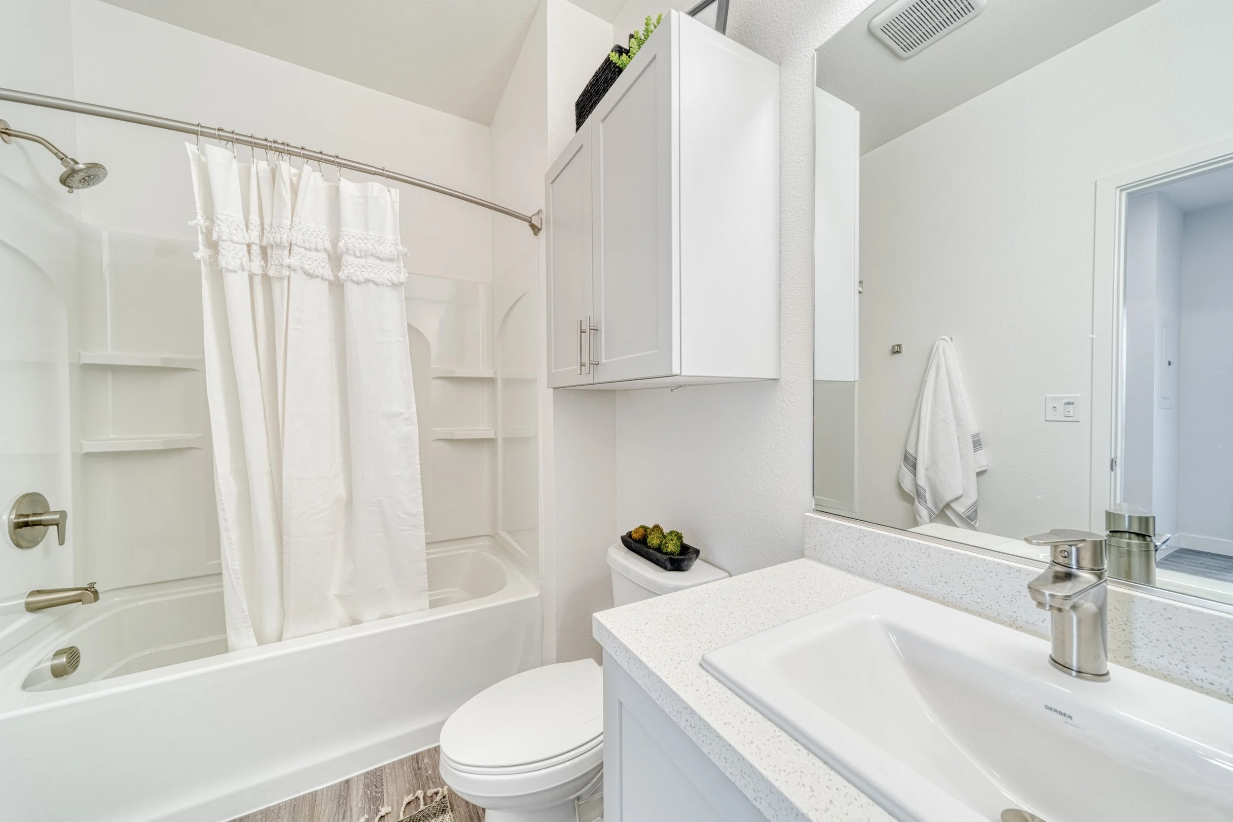 A small, modern bathroom with a white bathtub and shower, a countertop with a built-in sink, a large mirror, white cabinetry, and a toilet. The bathroom has white walls and a wood-style floor.