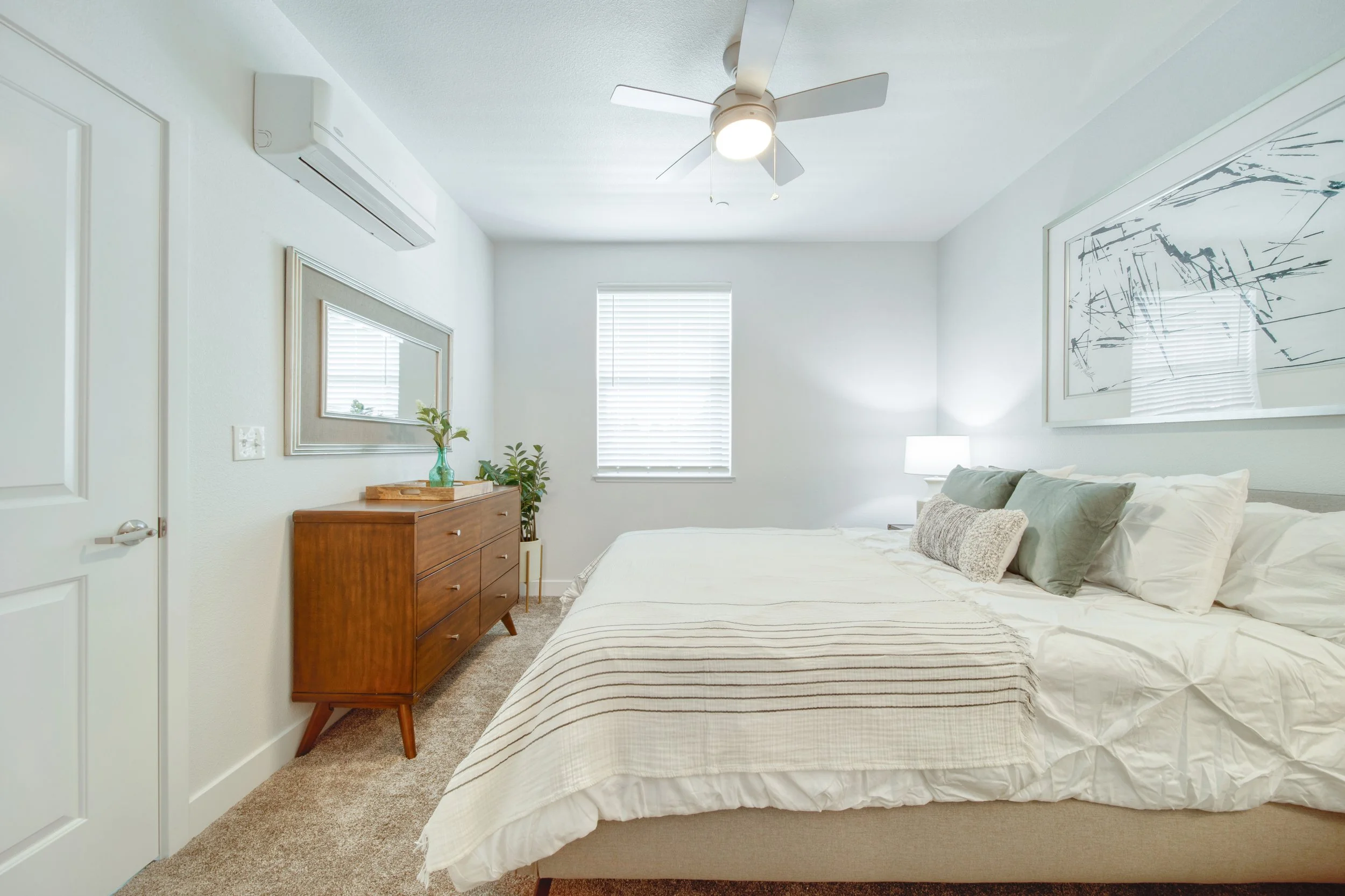 A bedroom with a bed, a wooden dresser, a mirror, a window with blinds, a ceiling fan, a lamp, a wall art piece, and some houseplants.
