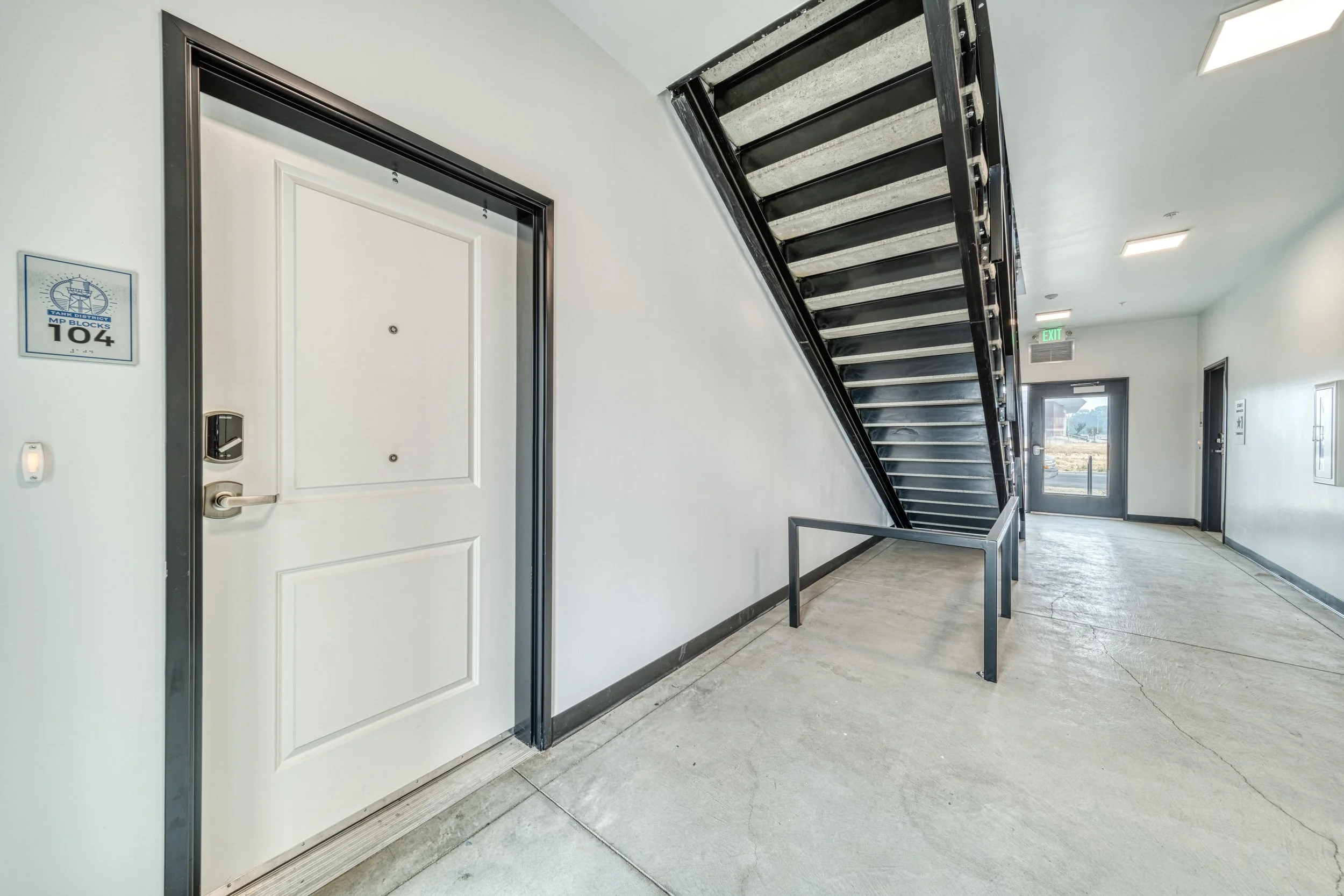 Interior view of an apartment or office building hallway featuring a white door, a staircase with black metal framing, an exit door at the end of the hallway, and a fire alarm panel on the wall.