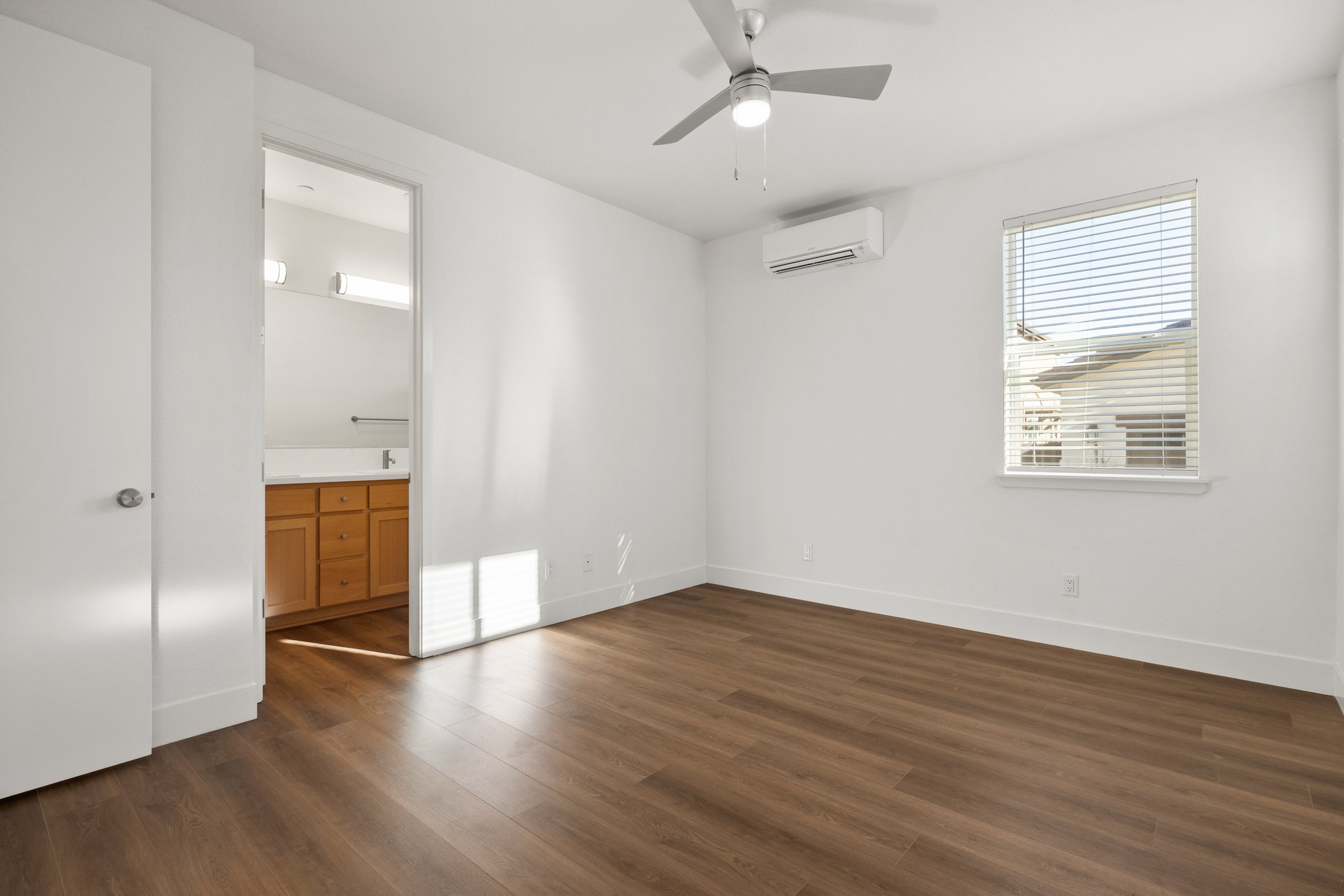 Empty bedroom with white walls, hardwood flooring, ceiling fan, window with blinds, and an attached bathroom with wooden cabinets.
