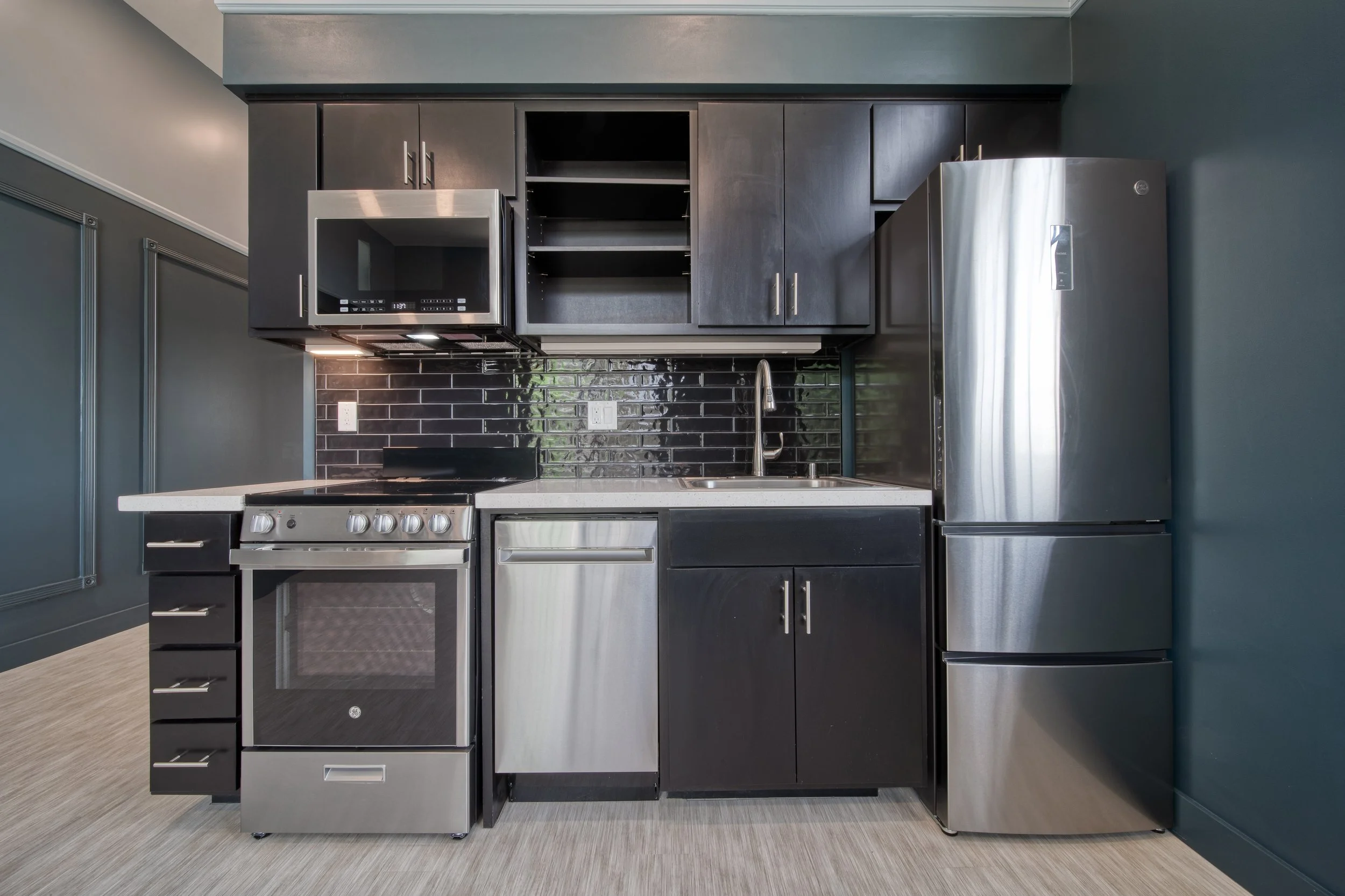 Modern kitchen with dark cabinets, stainless steel appliances including a refrigerator, oven, microwave, and dishwasher, black subway tile backsplash, white countertop, and a dark green wall.