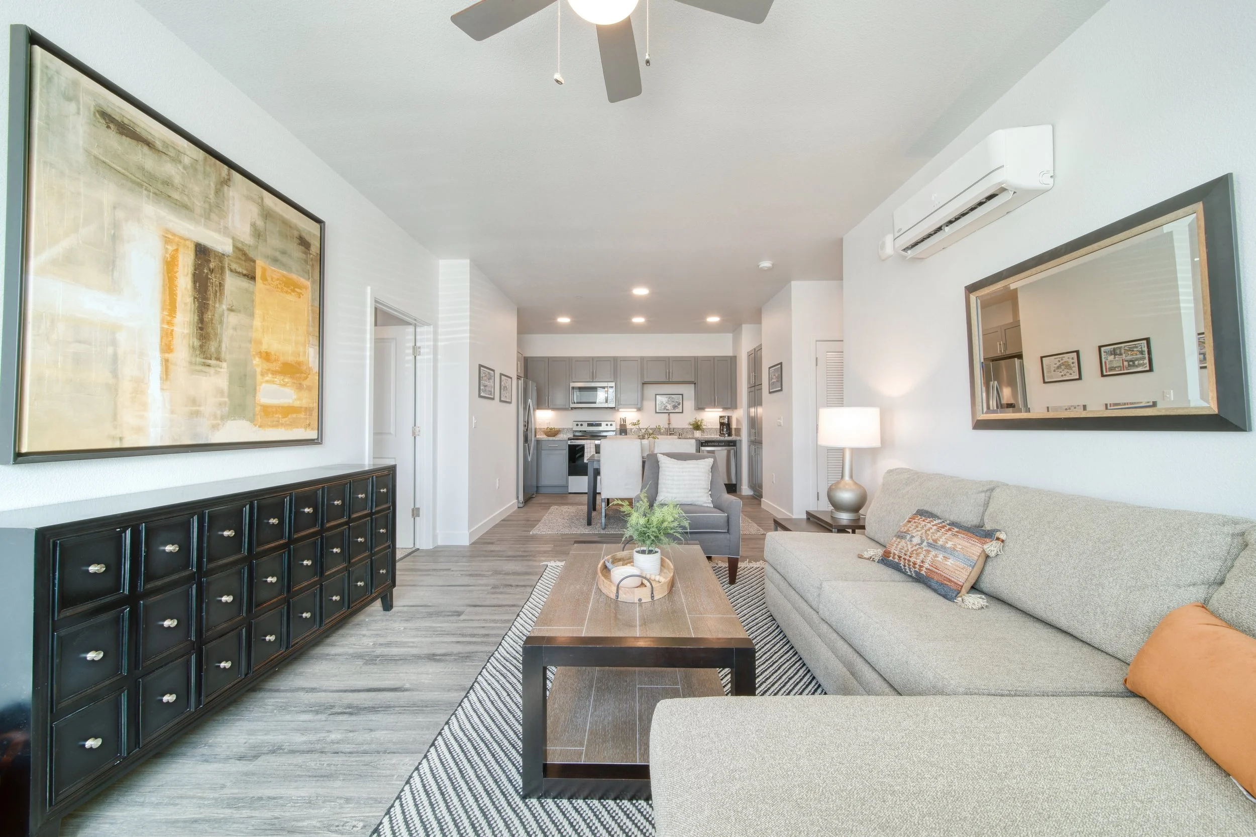 Living room with gray sofa, beige armchair, wooden coffee table, black sideboard, wall art, mirror, and kitchen in background.